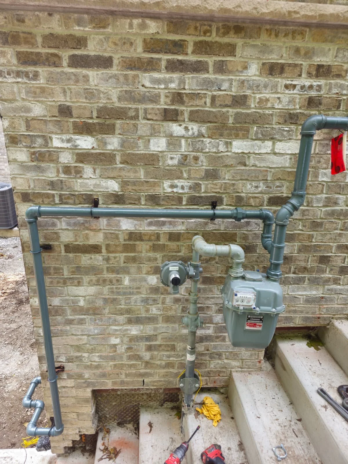 Gas meter and plumbing pipes on brick wall outside a house, with construction tools on concrete stairs.