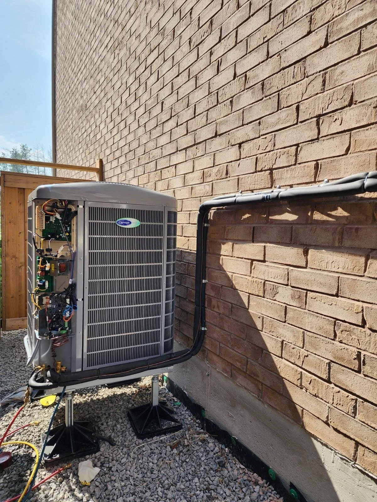 Outdoor air conditioning unit installed next to a brick wall, with electrical wiring and service pipes connected, on a gravel ground with a wooden fence in the background.
