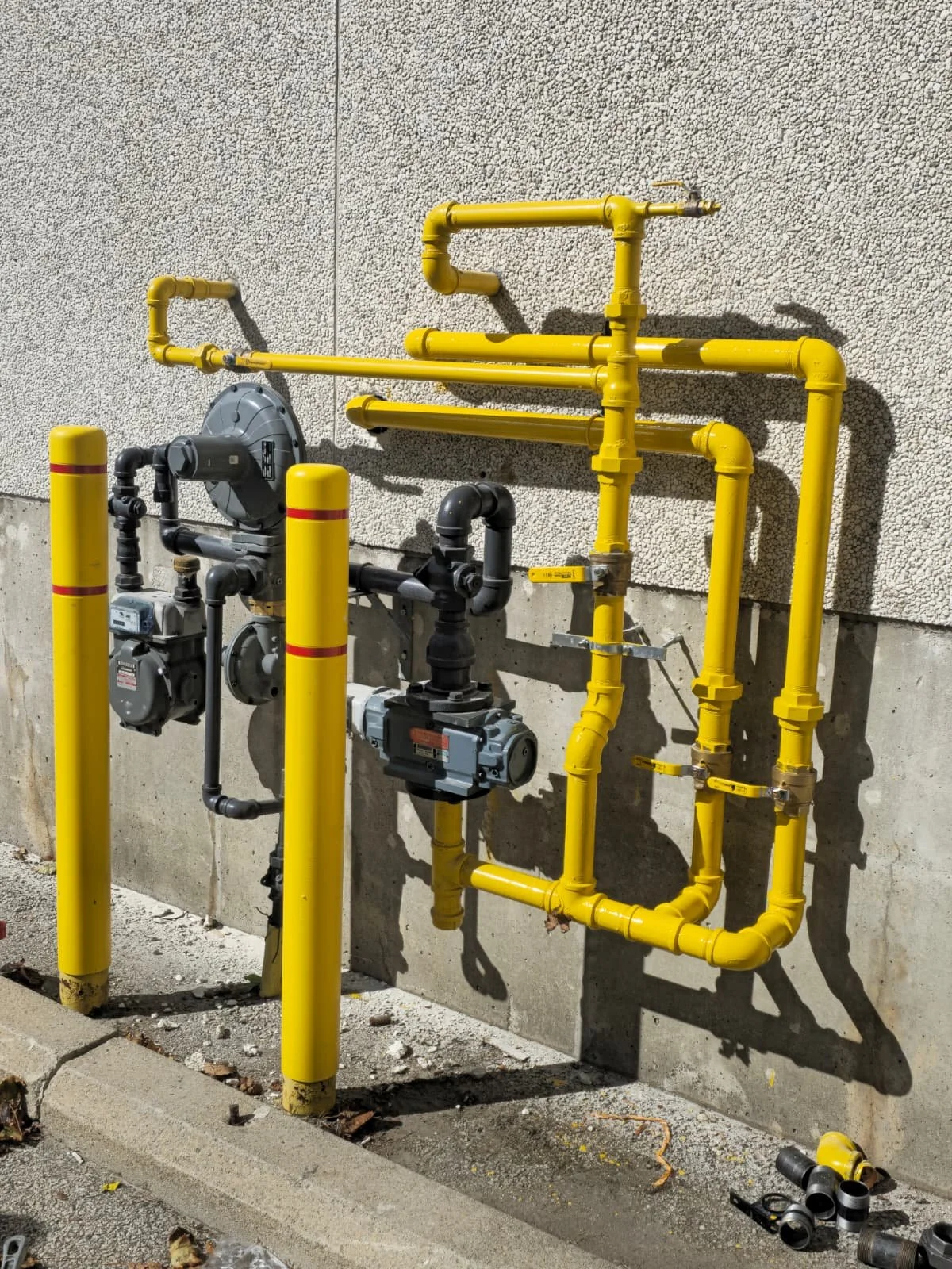 A network of yellow and black industrial pipes mounted on a concrete wall outside, with some valves and fittings attached.