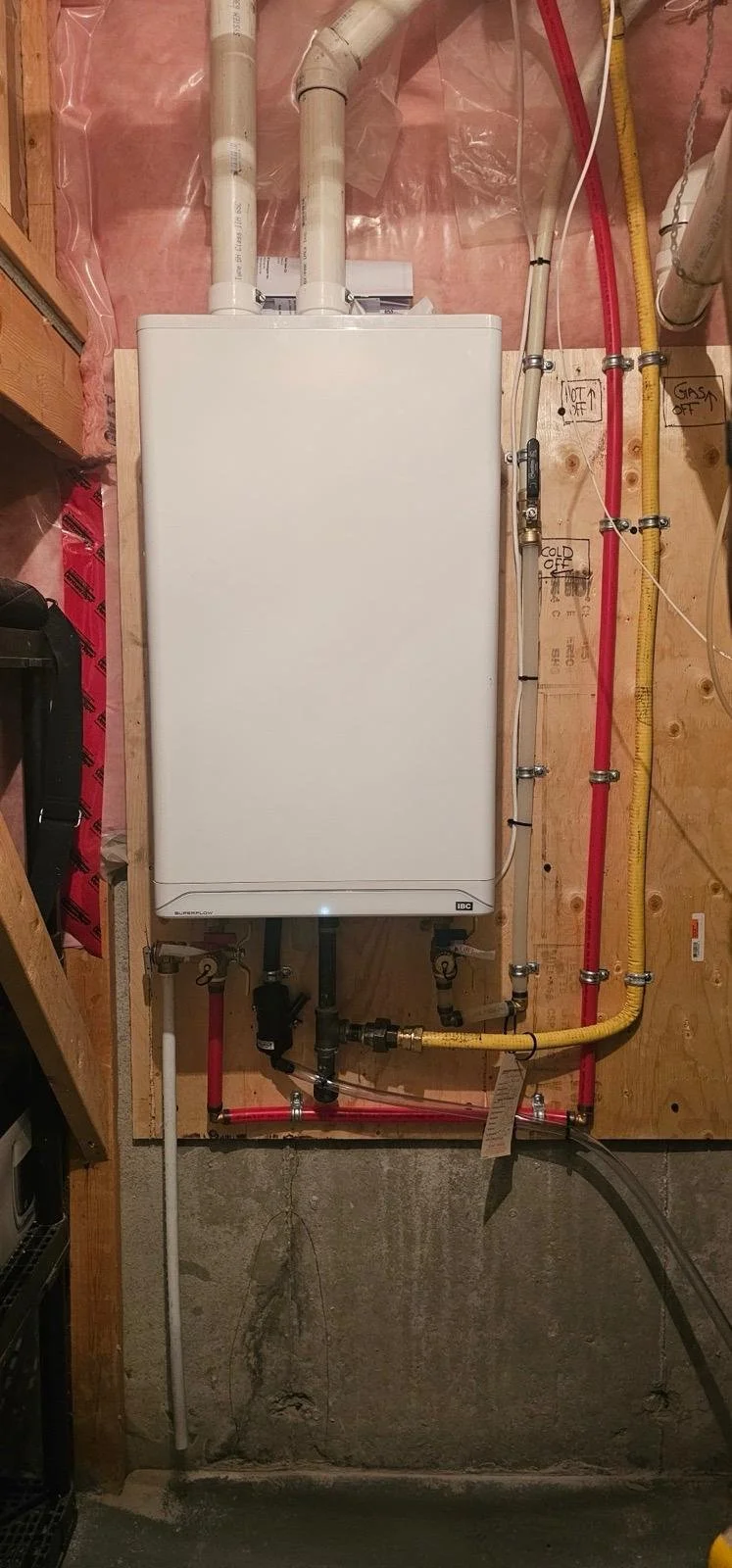 A water heater system installed on a wooden wall with multiple pipes and control labels, including hot and cold water lines, and an electrical conduit.