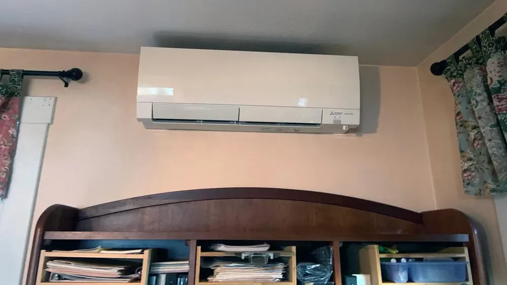 A split air conditioning unit mounted on a beige wall above a wooden bookshelf filled with books and papers, with floral curtains on both sides.
