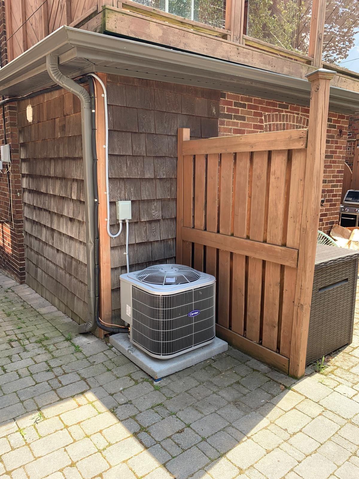 Outdoor air conditioning unit next to a wooden privacy fence, on a concrete pad with brick pavers.