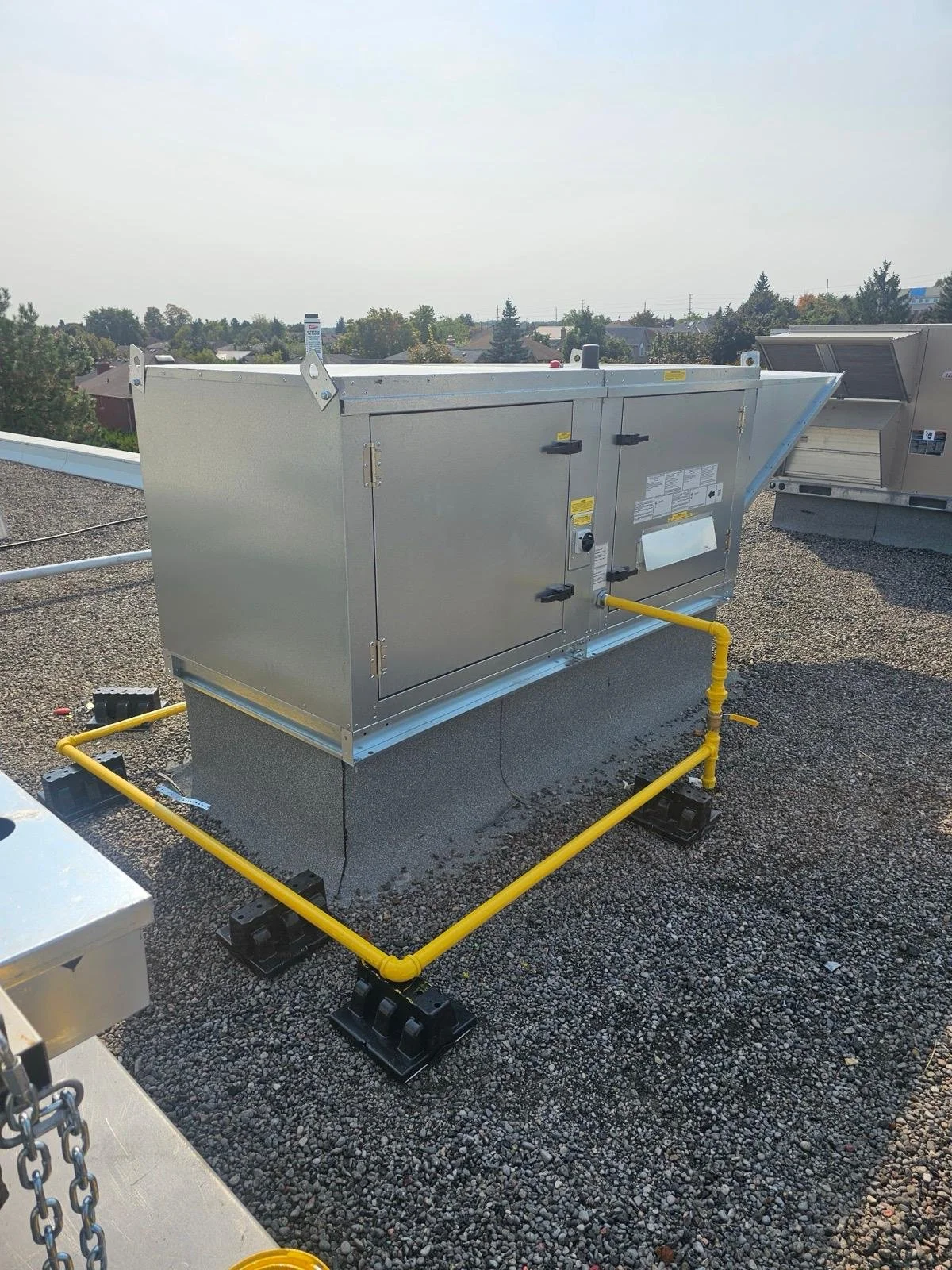 HVAC rooftop air handling unit installed on a gravel surface, surrounded by yellow piping and black supports, with a background of trees and a partly cloudy sky.