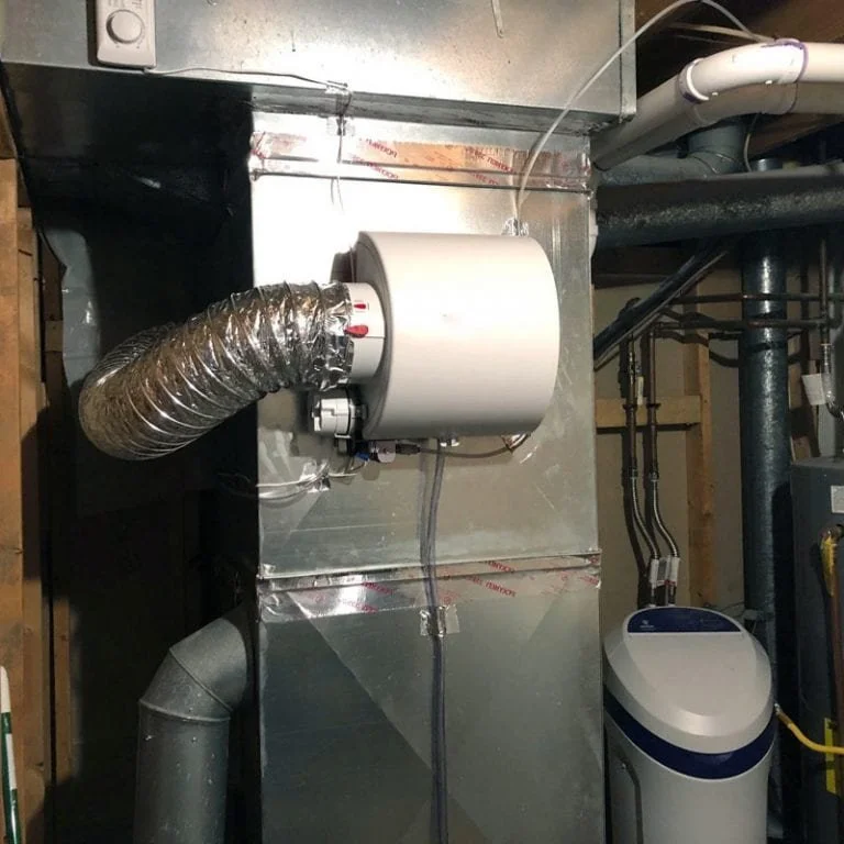 A furnace with a flexible metal duct connected to it, installed in a basement or utility area with visible pipes and electrical wiring.