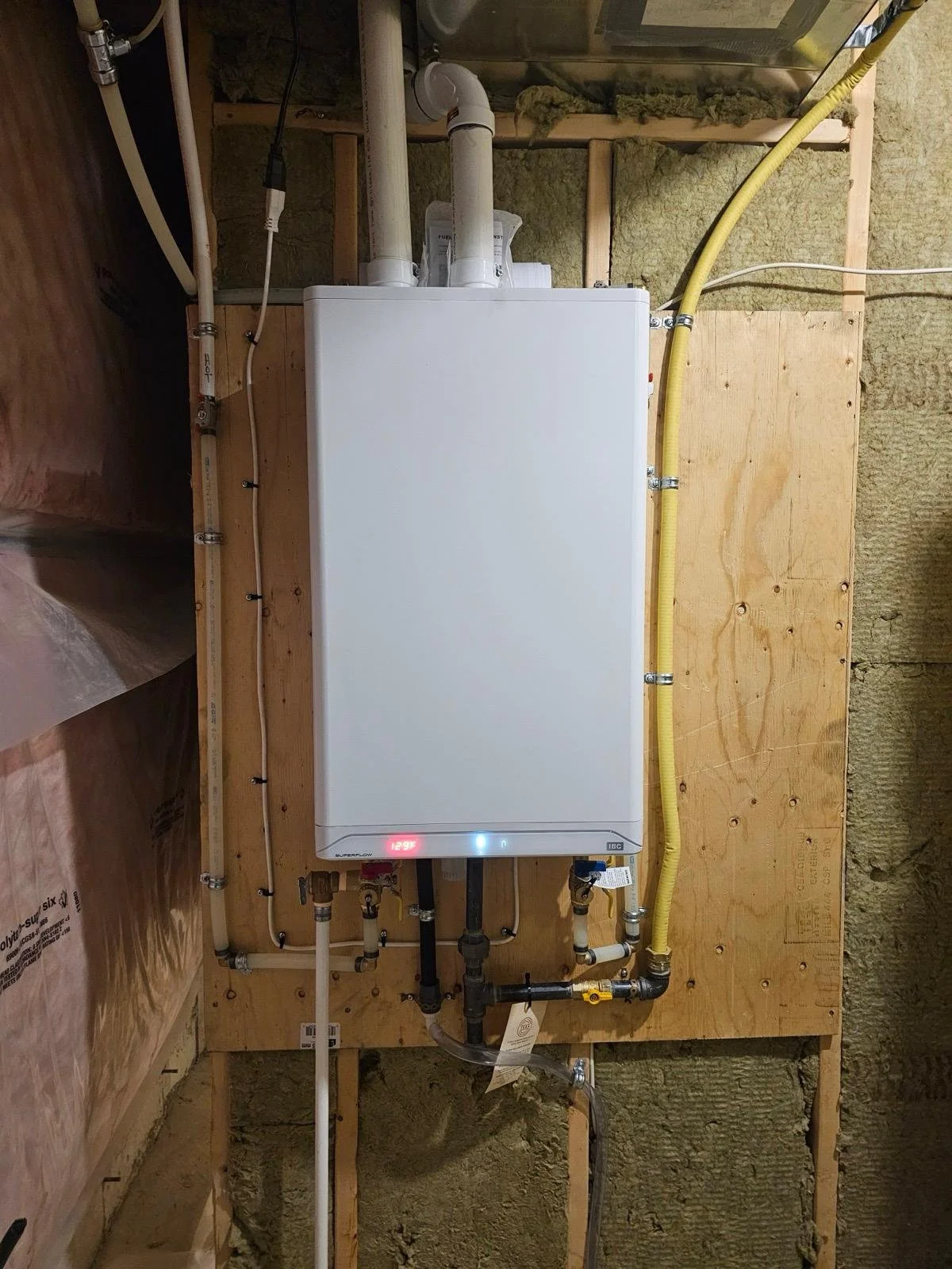 A tankless water heater installed on a wooden wall with pipes, ducts, and insulation around it.