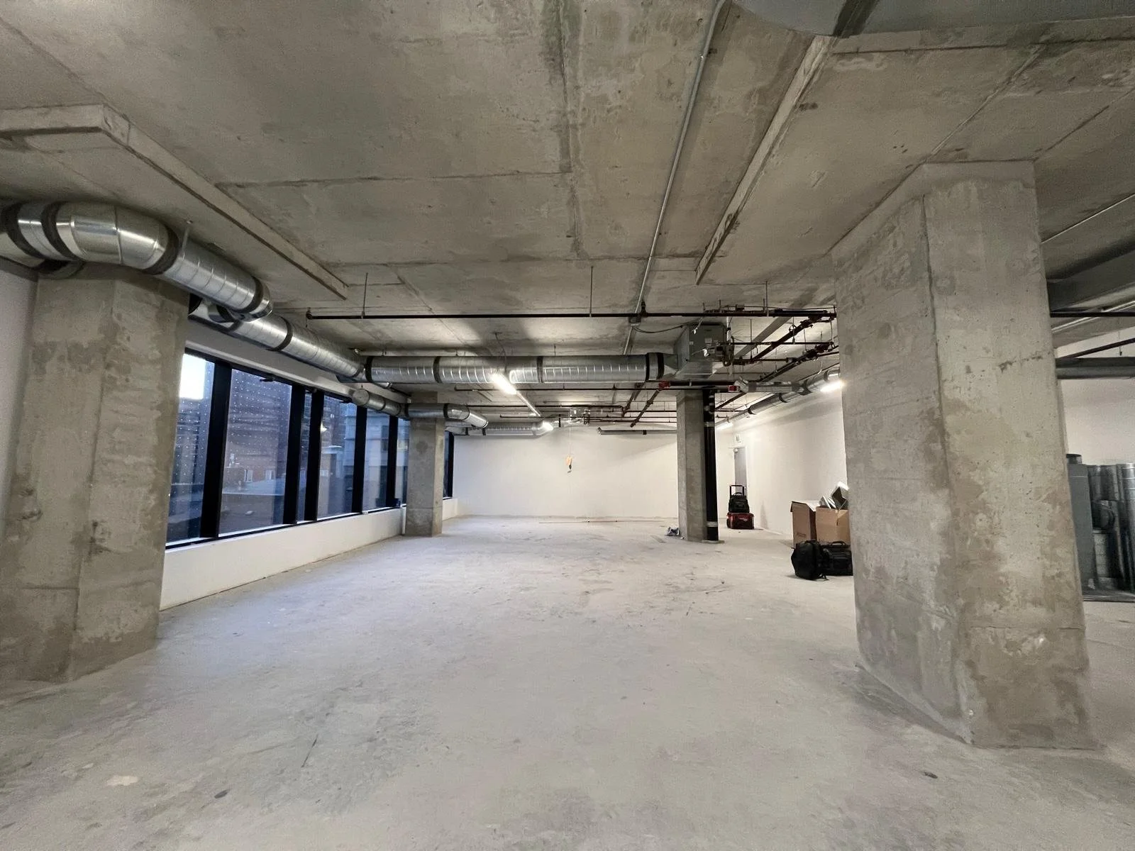 Empty, unfinished interior space under construction with concrete floors, large windows, visible ductwork, pipes, and construction tools.