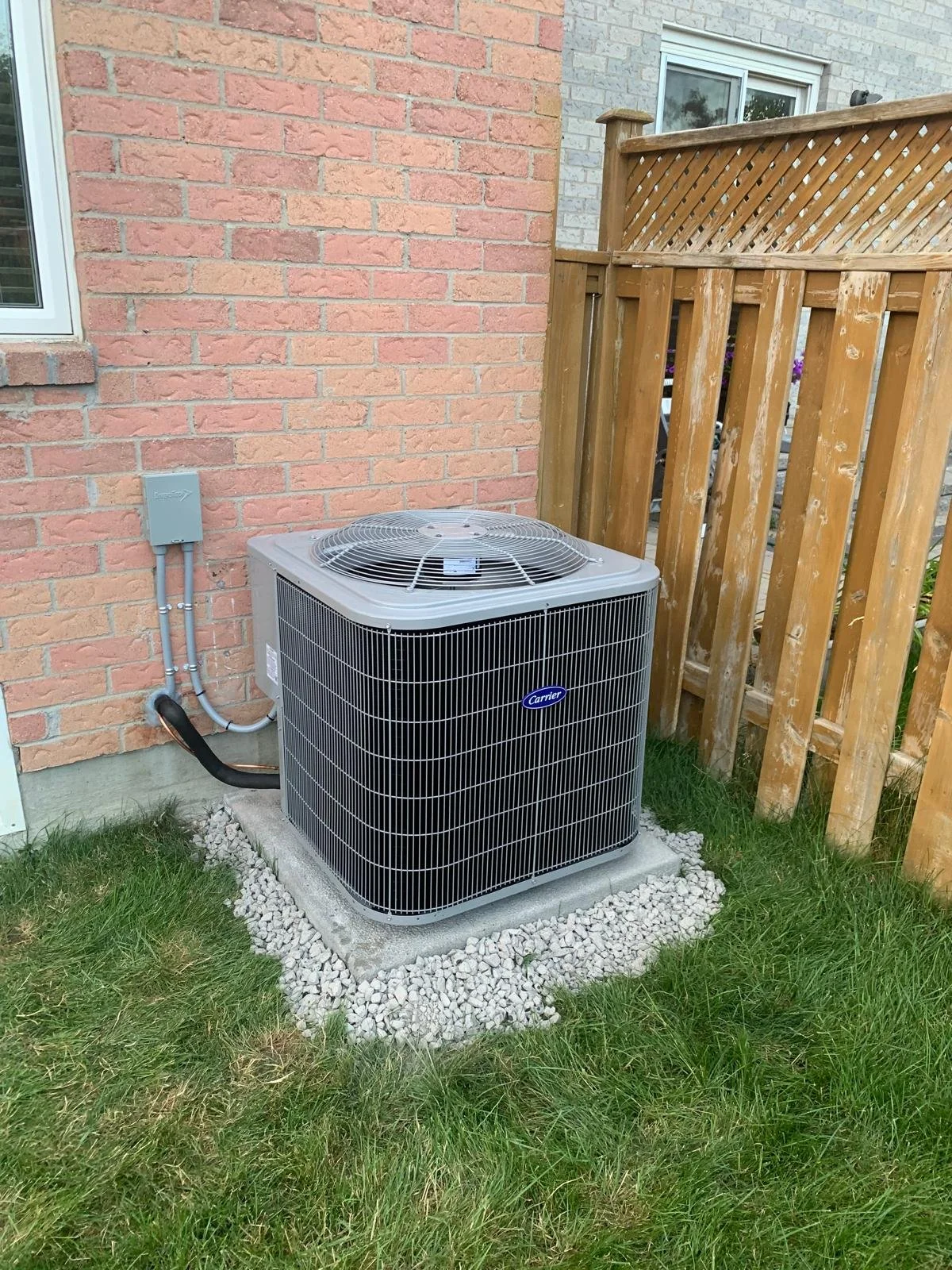 Outdoor air conditioning unit on a concrete pad next to a brick house wall and a wooden fence, with grass in the foreground.