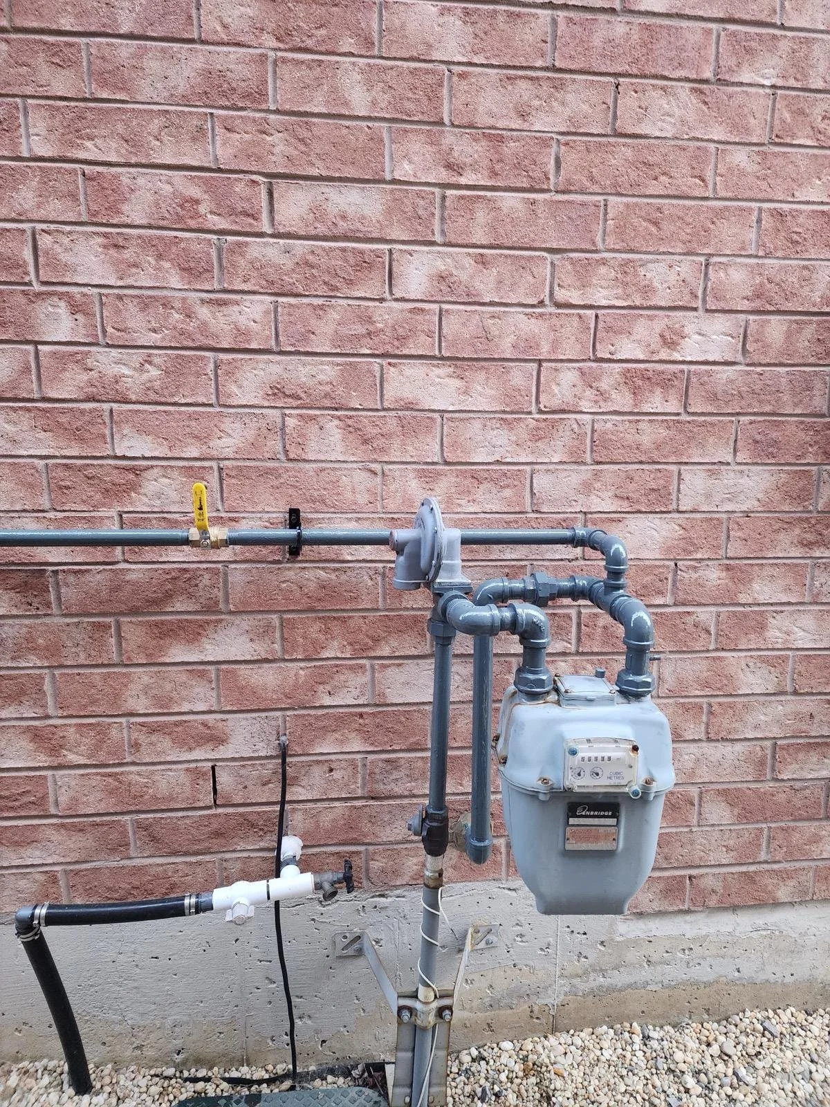 Outdoor view of a gas meter connected to gas pipes against a brick wall with gravel on the ground.