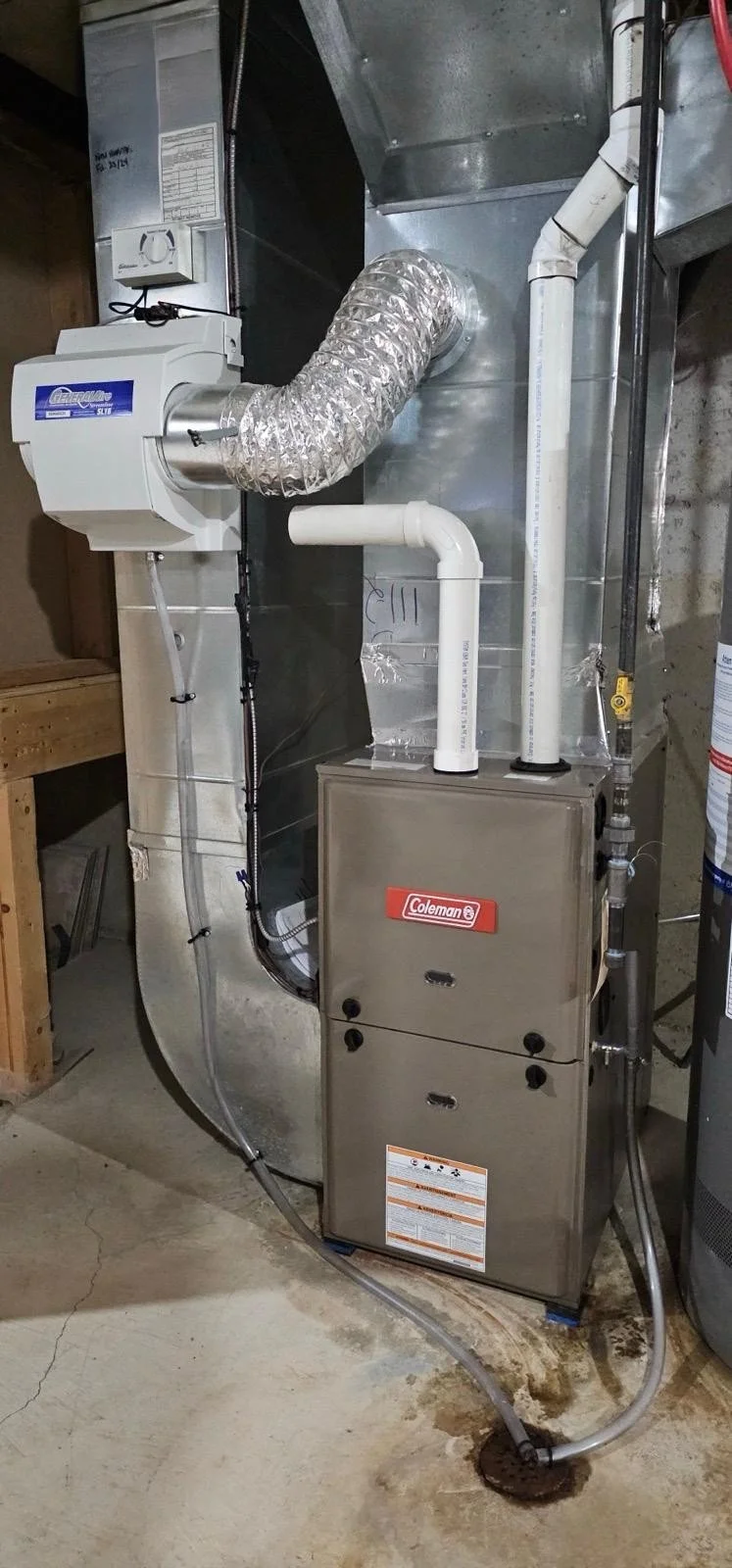 A furnace and duct system in a basement or utility room with PVC pipes, metal ducts, and electrical wiring.