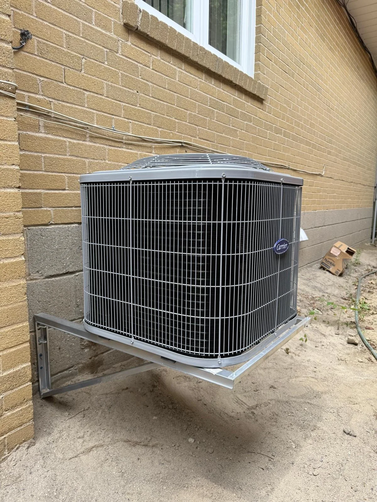 An outdoor air conditioning unit mounted on a metal stand beside a brick house wall.