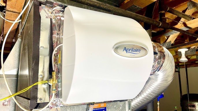 A white Aprilaire humidifier mounted on a ceiling, connected to ventilation ductwork with metal and flexible hose in a utility room.