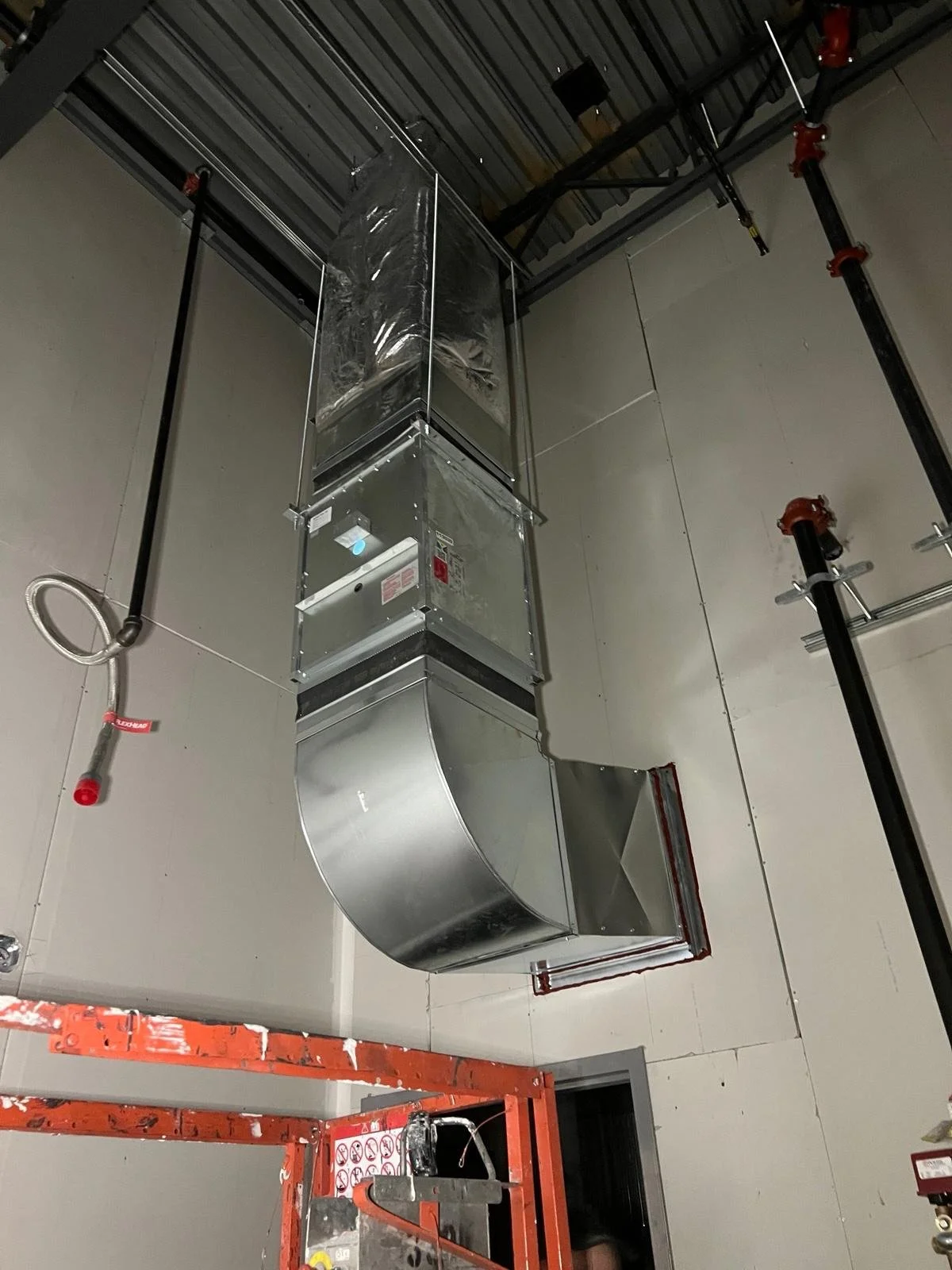 An unfinished ceiling with industrial air conditioning ductwork and PVC pipes, partially covered in plastic wrap, in a construction site.