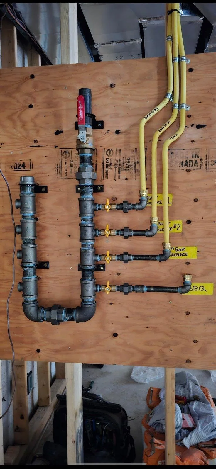 A plumbing piping system with multiple pipes and valves on a plywood wall, labeled with yellow tags indicating different function areas such as appliance #2, floor replacement, and BBQ.