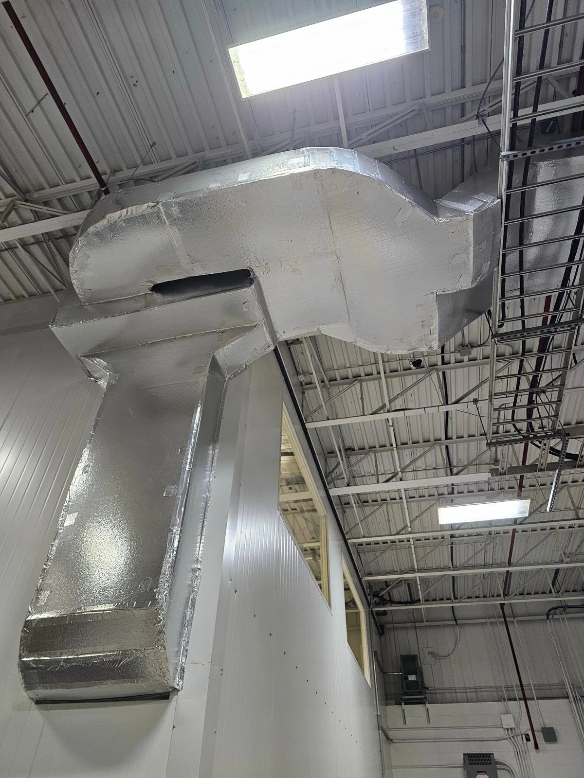 Large metallic ductwork installed on the ceiling of an industrial or commercial building with exposed ceiling beams and fluorescent lighting.