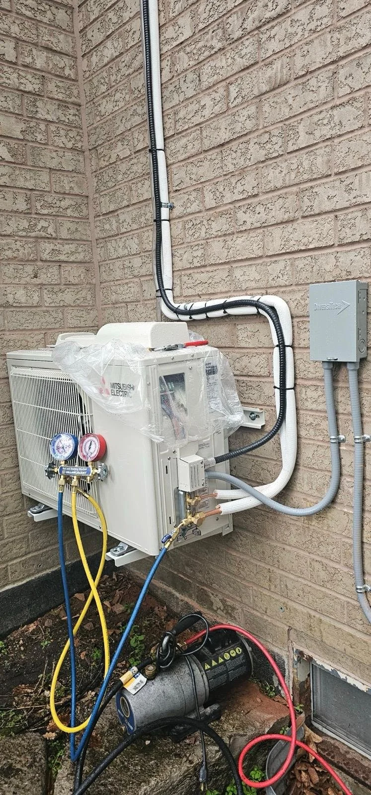 An outdoor HVAC unit connected to electrical wiring, refrigerant gauges, and a vacuum pump on a brick wall.