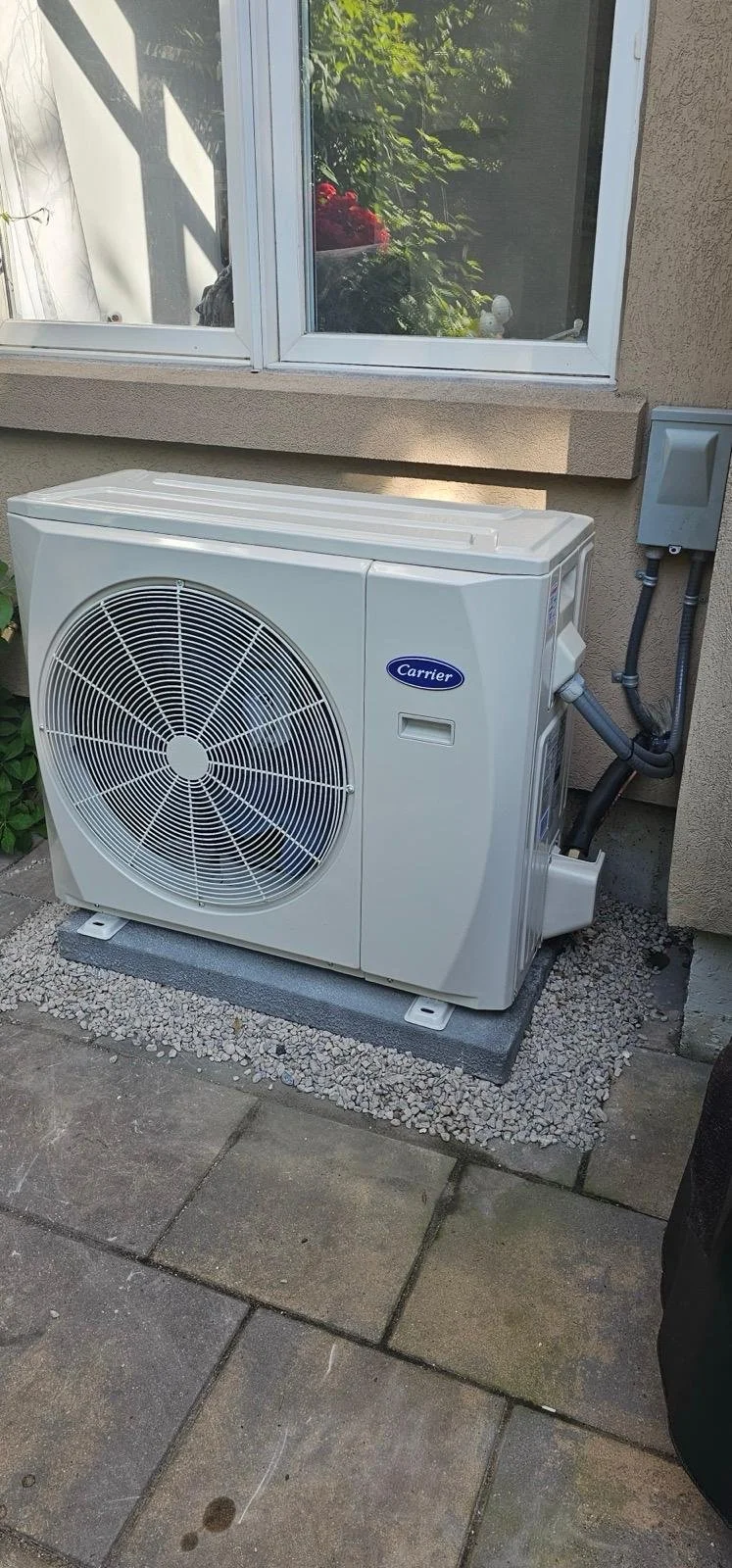 Outdoor air conditioning unit outside a house, positioned on a concrete slab with gravel, next to a window and beige wall.