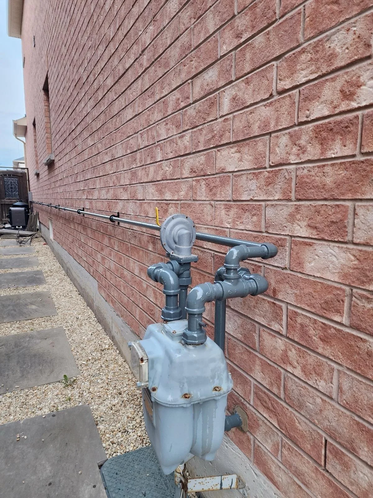 Exterior view of a brick building with a gas meter and pipe installation along the side, with concrete paving stones and gravel on the ground.