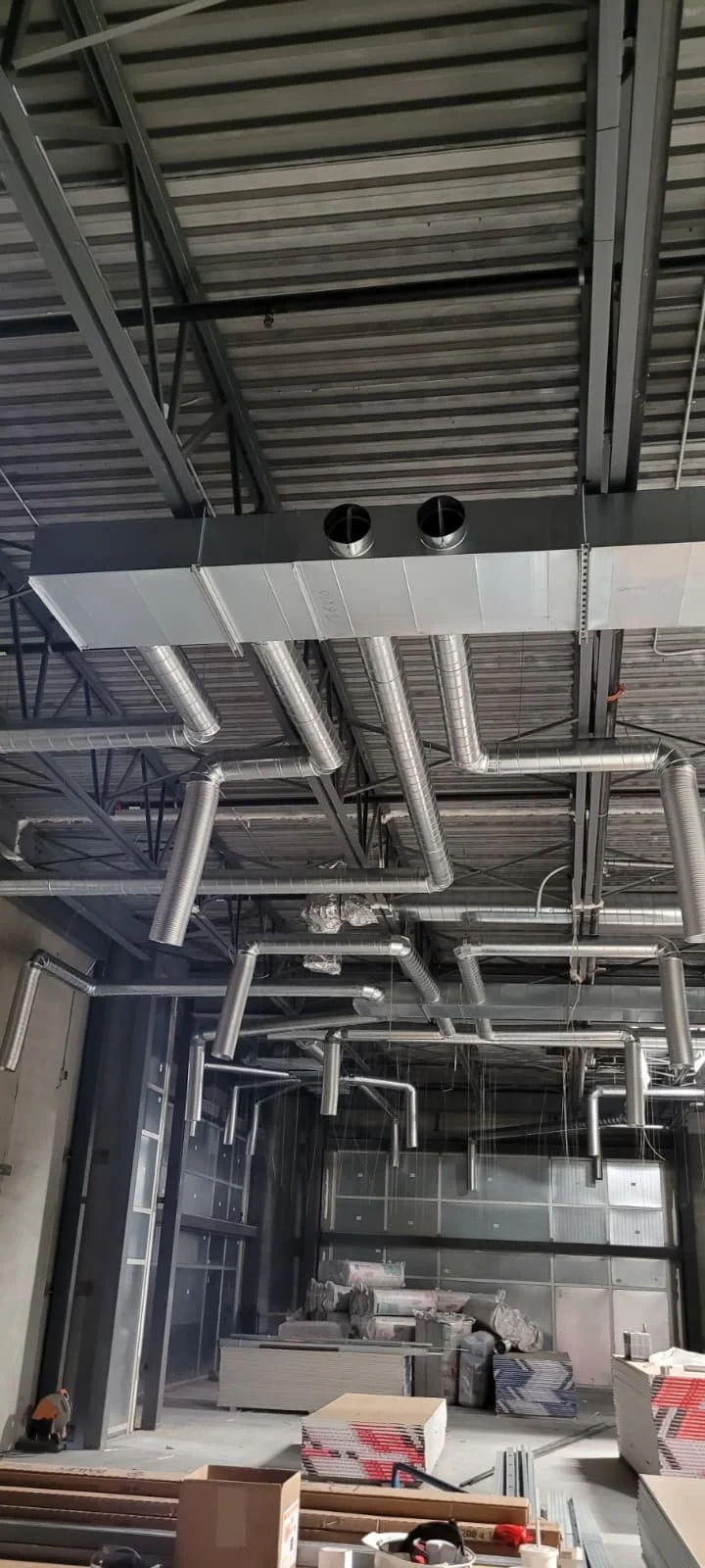 Interior view of a construction site with exposed metal ducts and pipes suspended from the ceiling, and construction materials and equipment on the floor.