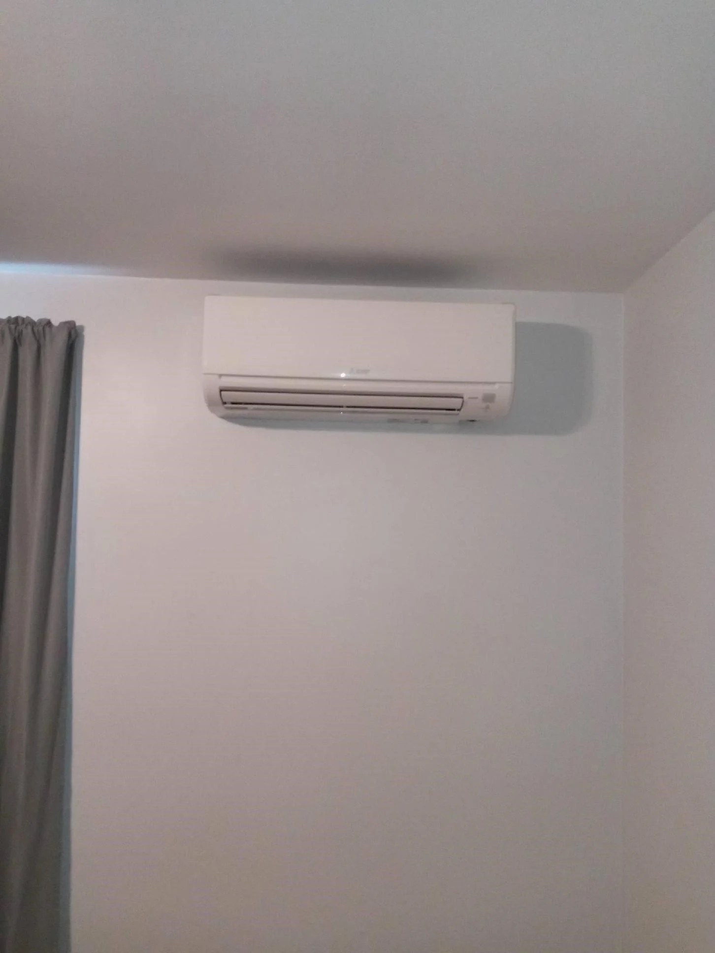 White wall-mounted air conditioner unit on a plain white wall, with gray curtains and a portion of a gray ceiling visible.