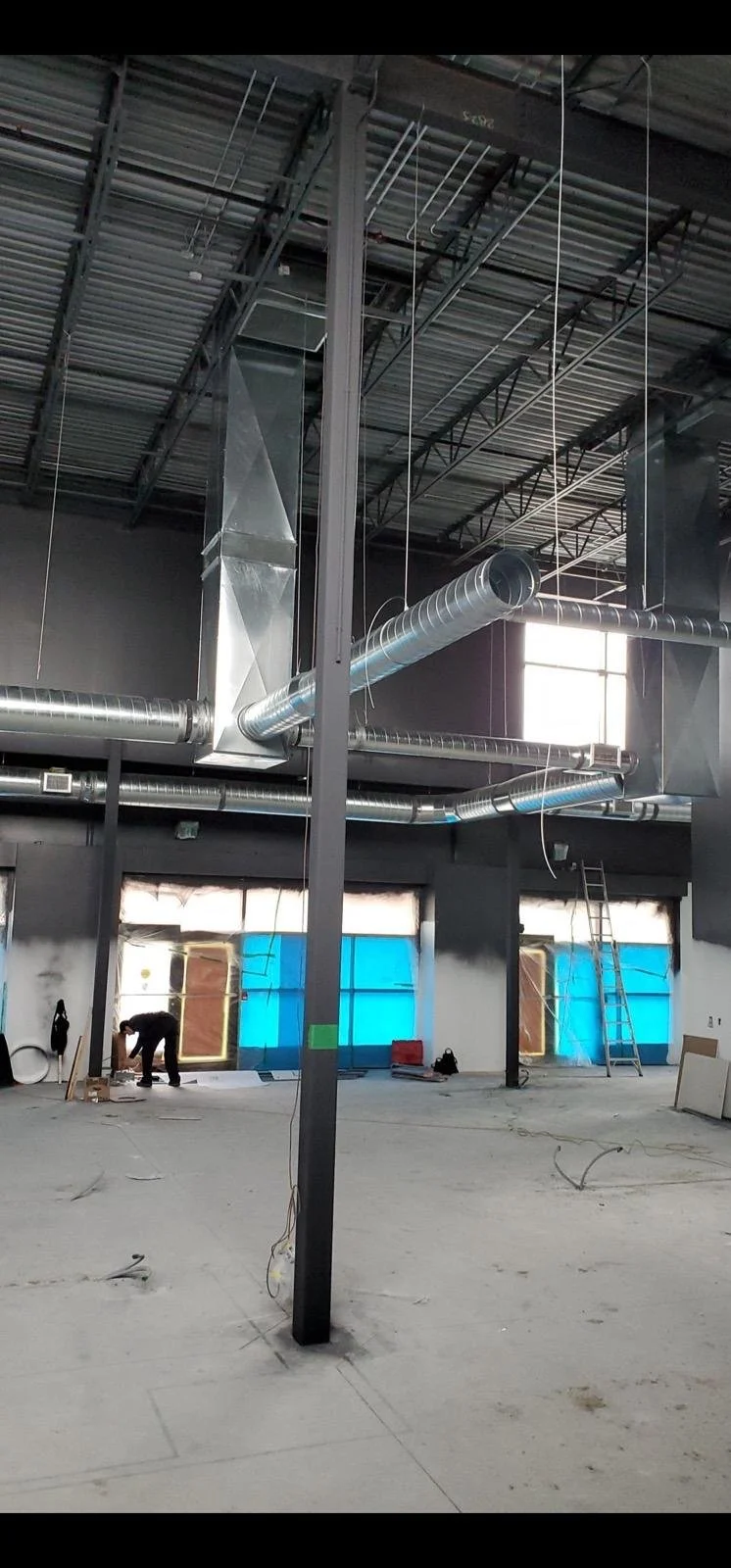 Interior of a building under construction with exposed ceiling, ductwork, and workers installing or finishing work.