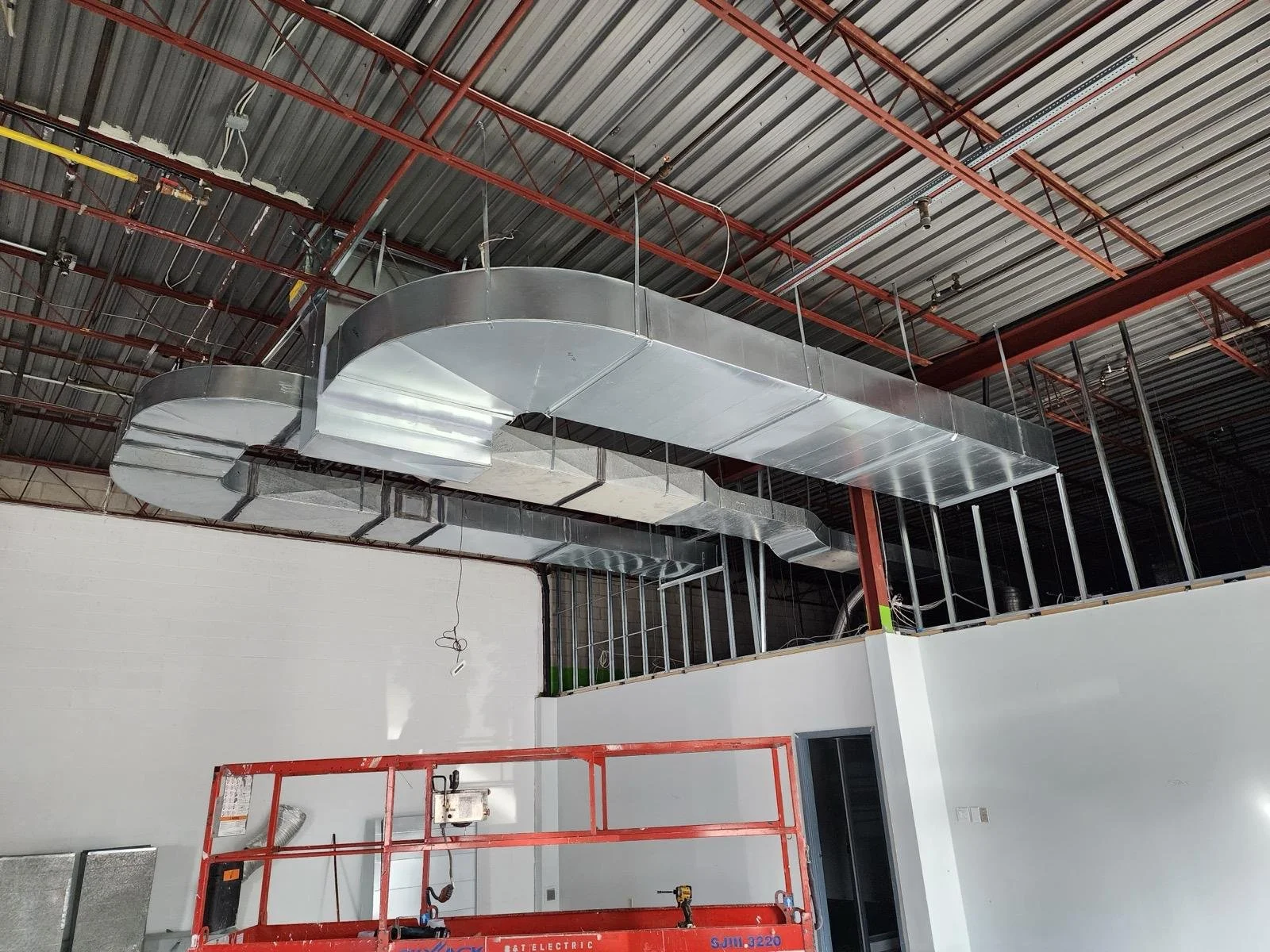 Construction interior with metallic air ducts, white walls, red scaffolding, and exposed ceiling pipes.