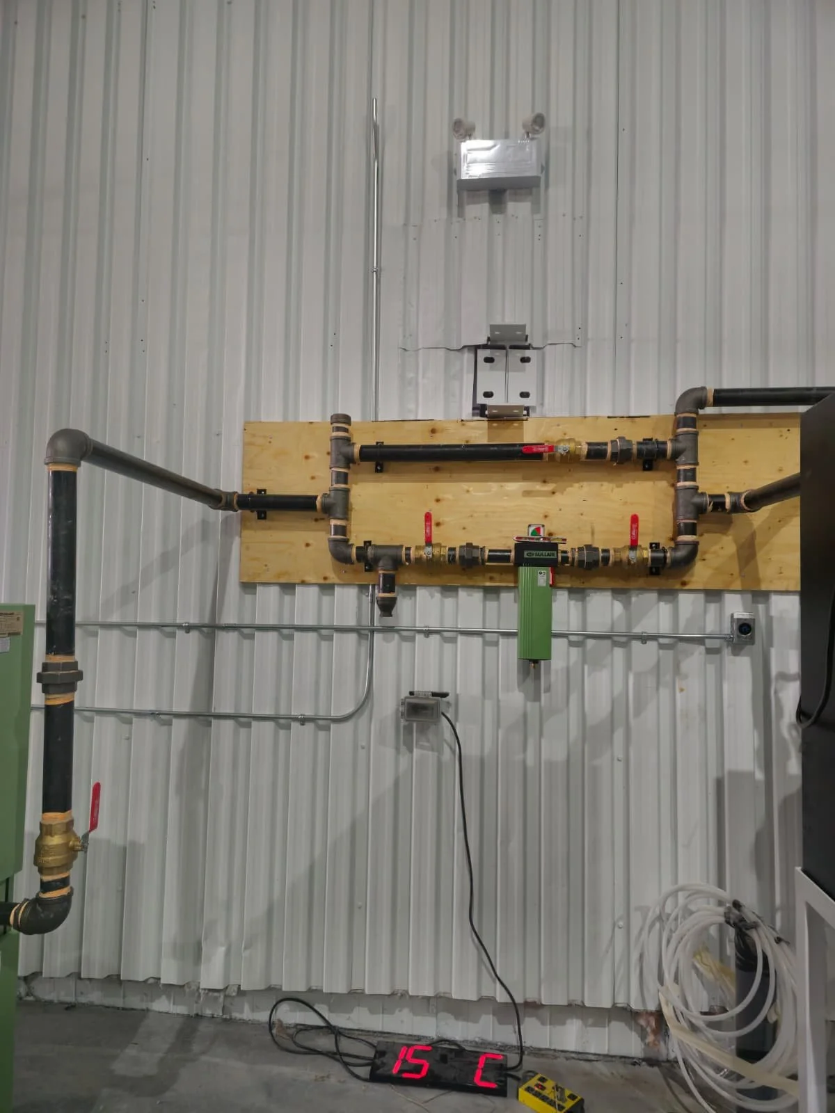 Industrial pipe system mounted on a wall with a digital temperature display reading 15°C on the floor in front of it.