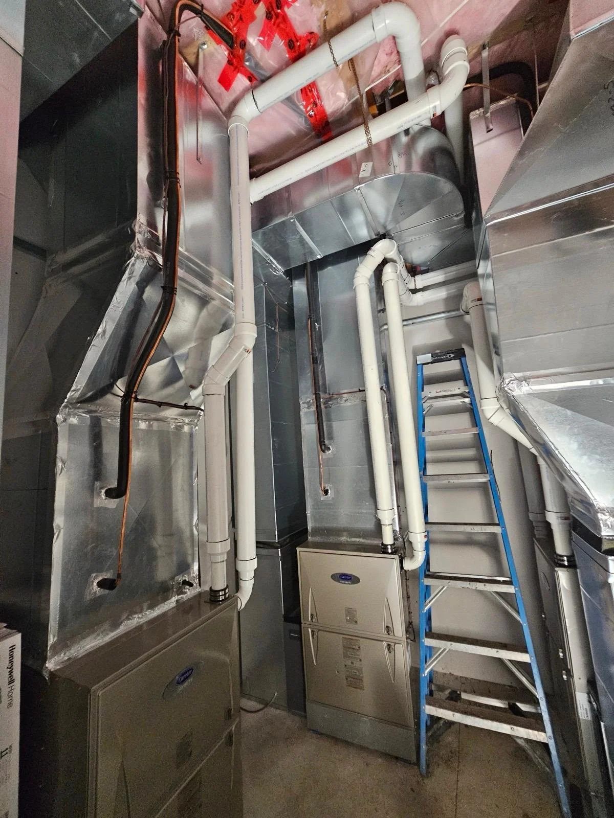 A utility or mechanical room with pipes, ducts, and a blue ladder. PVC pipes and metal ductwork run along the ceiling and walls, with some sections taped or insulated. There are two gray utility boxes on the floor.