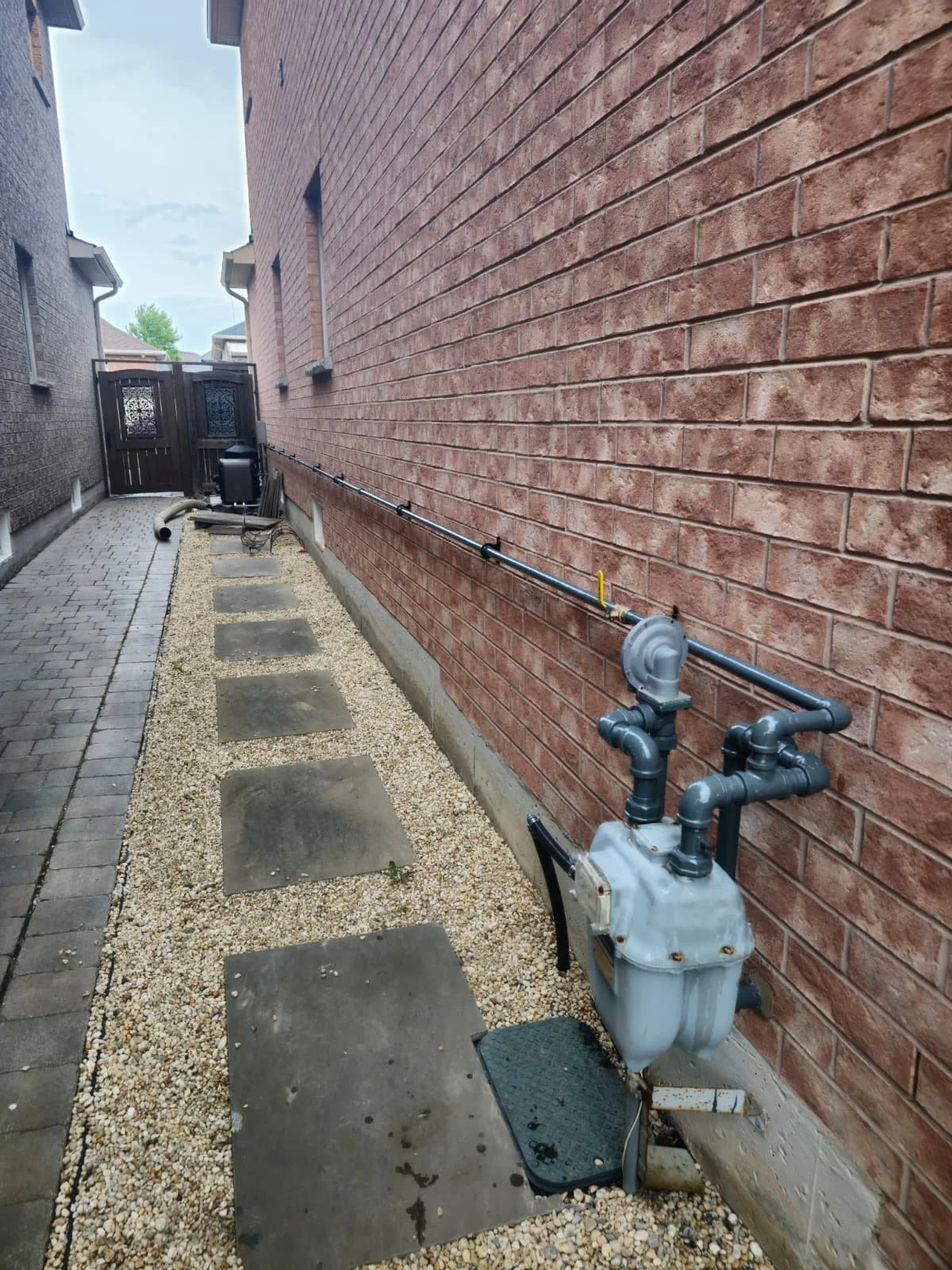 View of a narrow side yard with a brick wall on the right, concrete stepping stones on gravel, and an outdoor gas meter box with piping along the wall, leading to a black gate at the end.