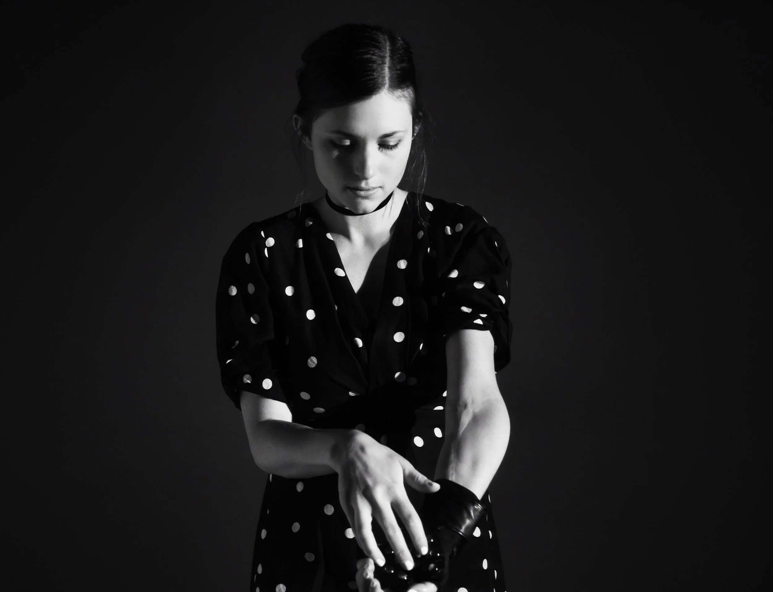 Molly is in a black polka-dot dress looking down, as she places a black prosthetic on her left arm. The photo is black and white, with soft lighting.
