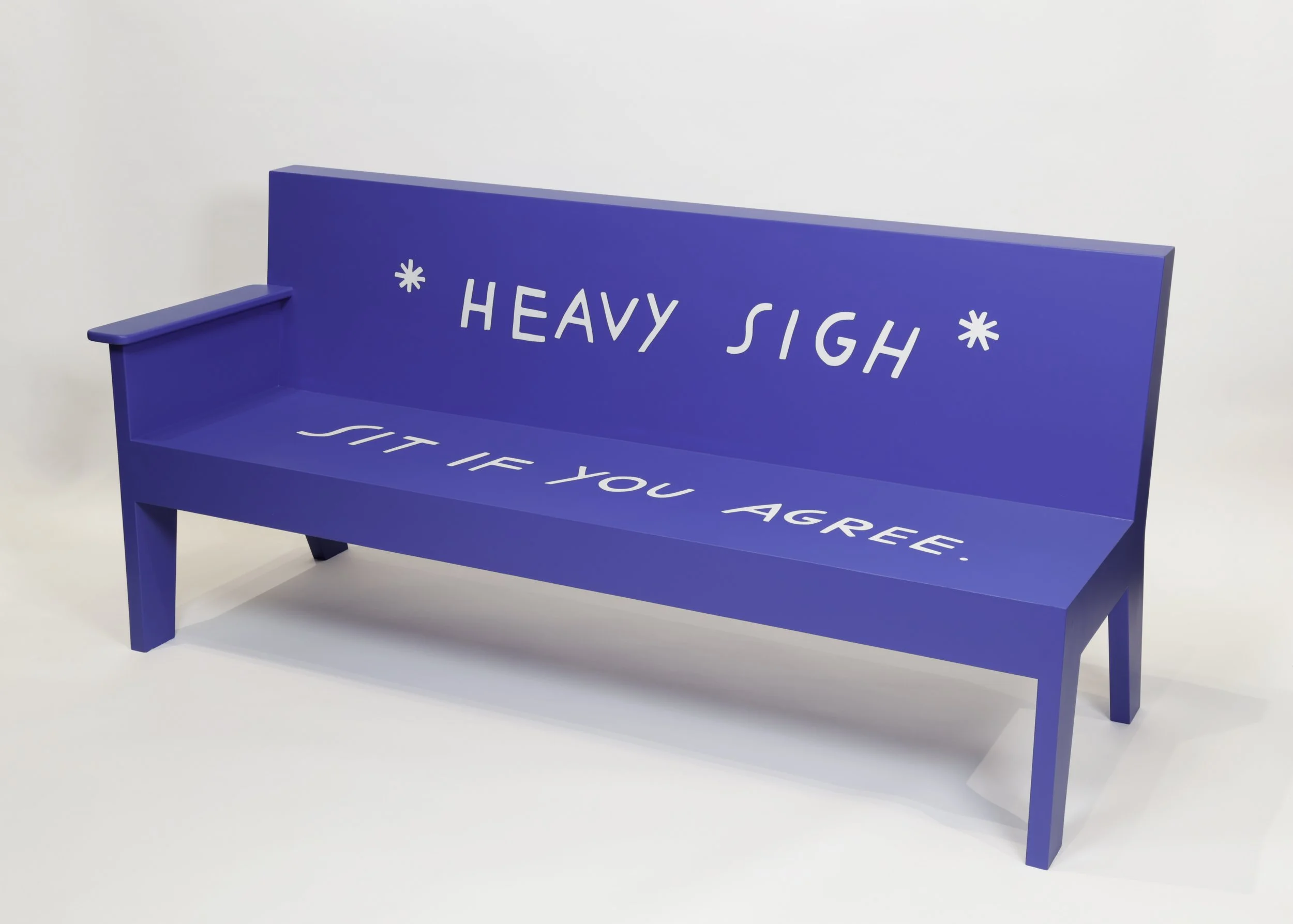 A simple wooden bench, with only one armrest, painted a shade of brilliant blue. On the back of the bench white letters spell “Heavy Sigh” within two asterisk marks. Continuing on the seat of the bench, the white letters read “Sit if you agree.”