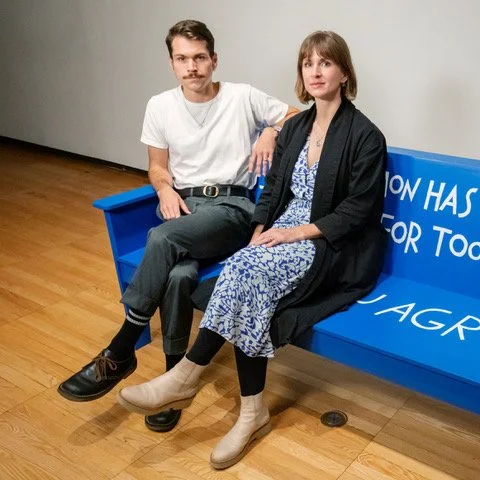 Christopher Robert Jones and Liza Sylvestre sit on a bright blue bench indoors.