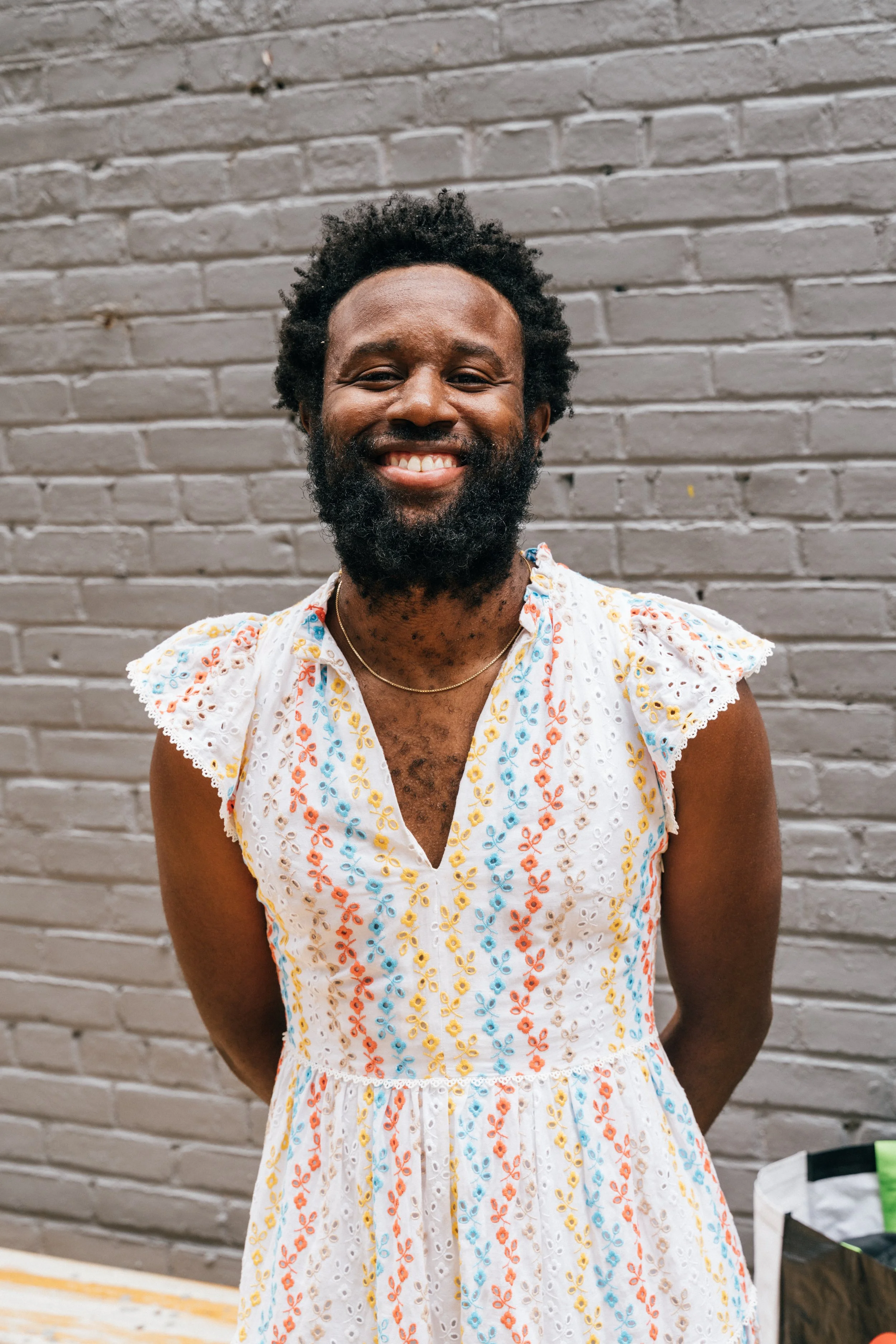 JJJJJJerome is smiling and wearing a white dress with colorful floral embroidery, standing in front of a gray brick wall.