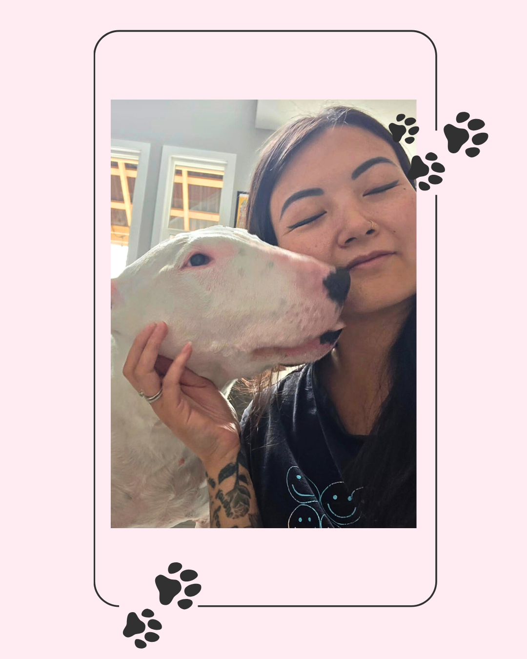 Woman with dark hair and tattoos holding a white Bull Terrier dog close to her face, both appearing happy indoors.