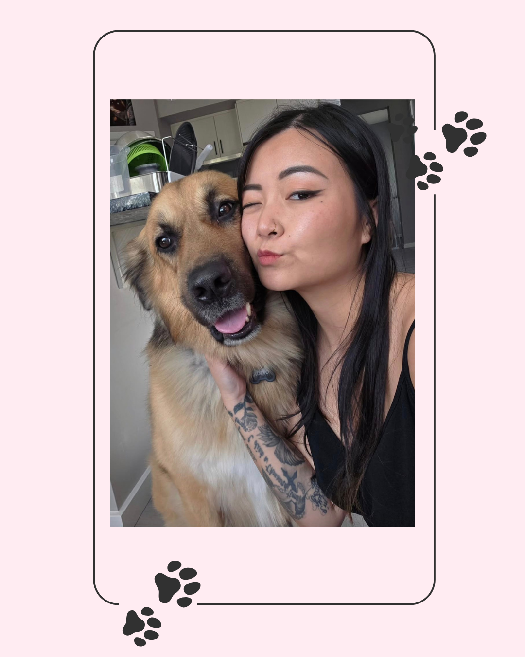 A woman with black hair and tattoos on her arm hugs a large, happy tan and black dog inside a home, with a kitchen in the background.