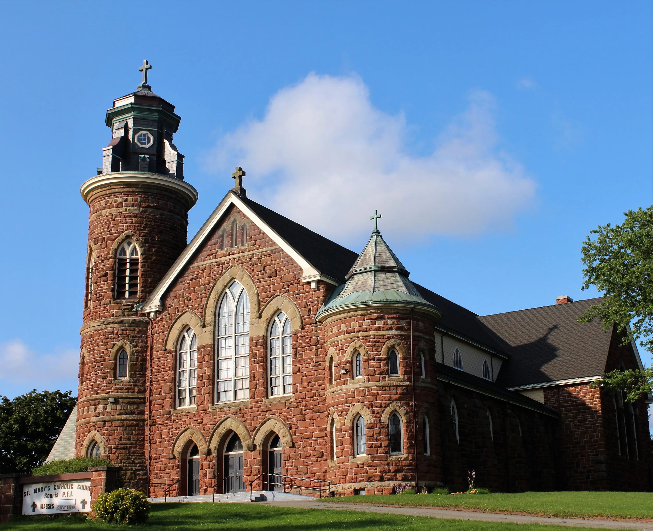 Red brick church with tall steeple, topped by a cross, and stained glass windows, set against a blue sky with clouds.