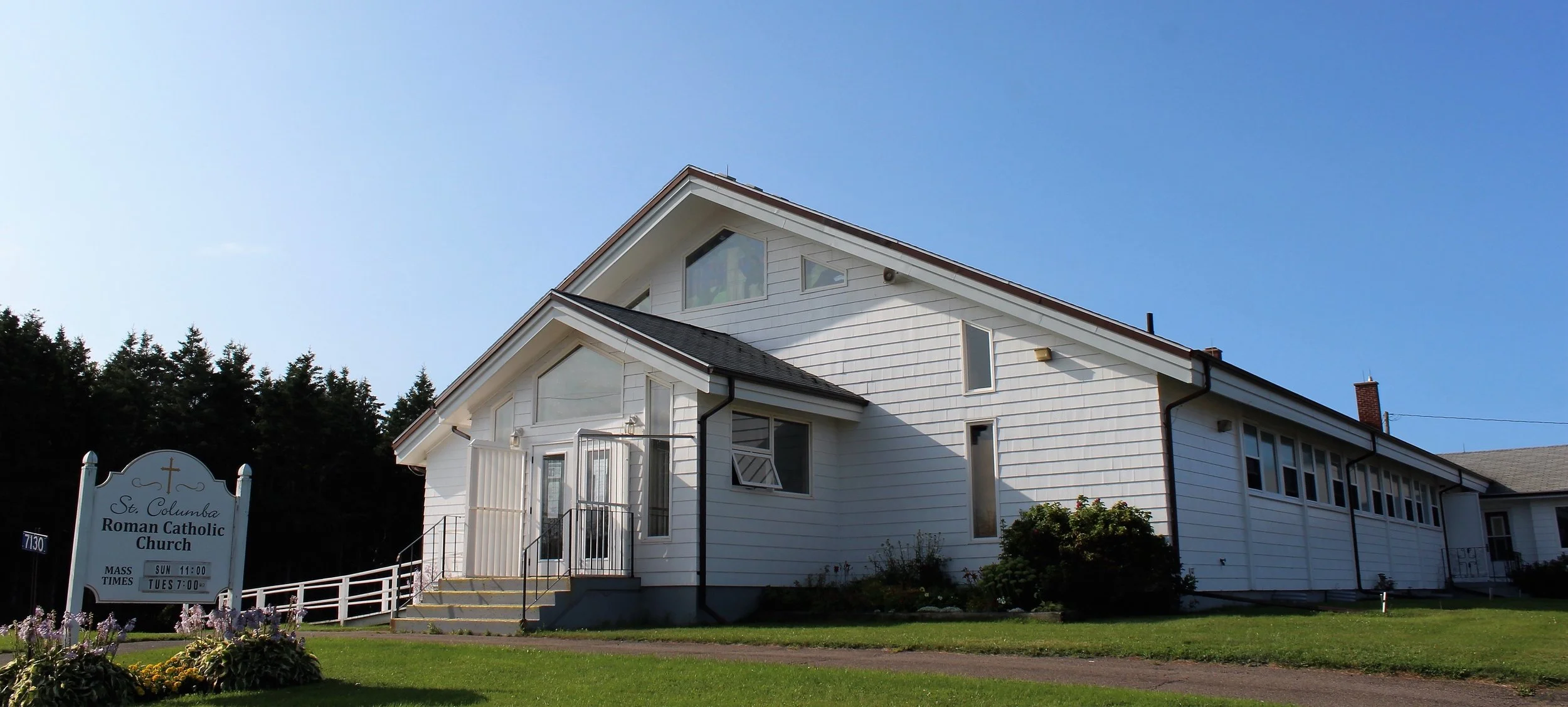 A white church building with a sign that reads 'St. Columba Roman Catholic Church' and service times, set in a grassy area with flowers. The church has a gabled roof, windows, and a small set of stairs leading to the entrance, with trees and a clear blue sky in the background.