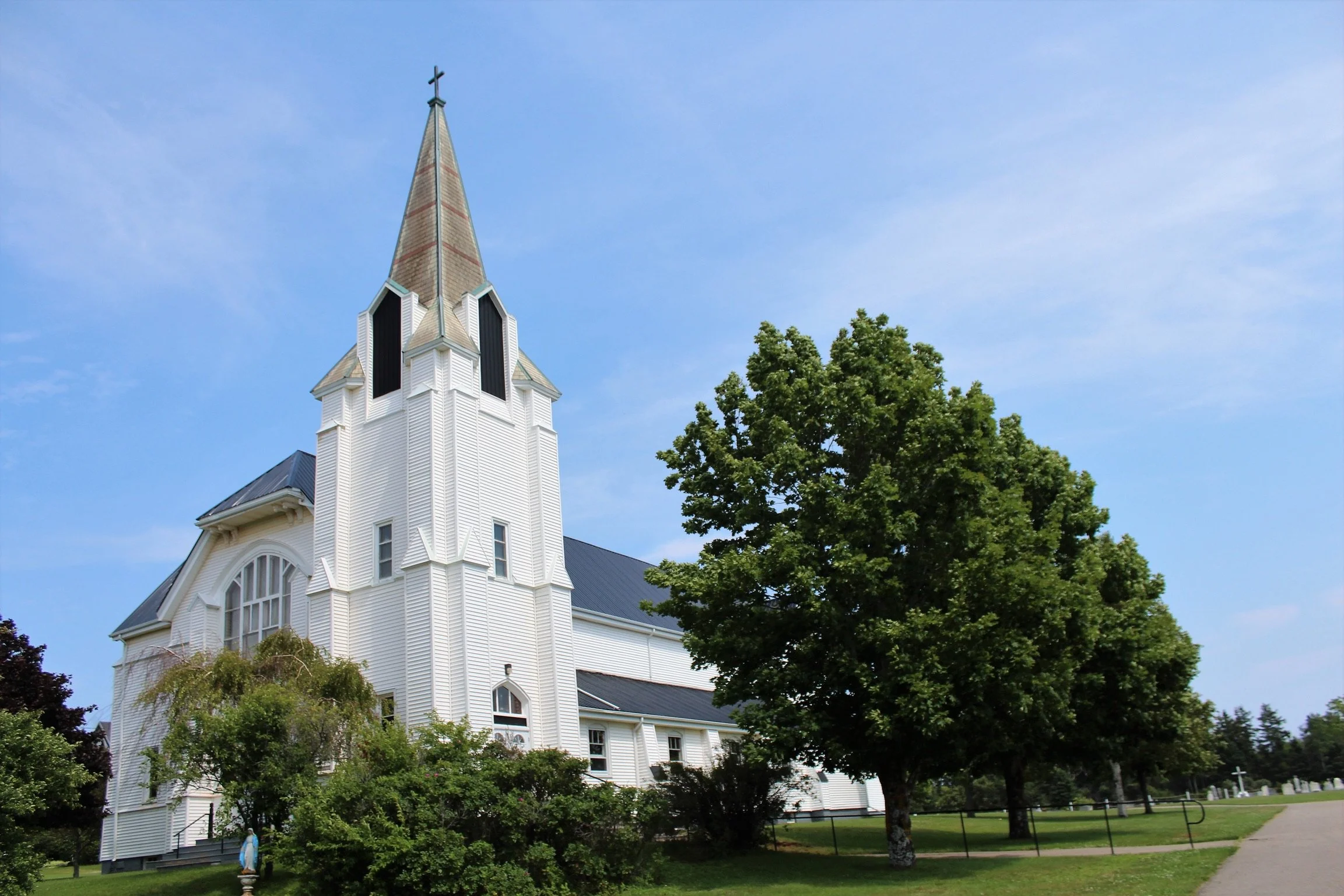 A white church with a tall steeple and cross on top, surrounded by green trees and grass, under a partly cloudy blue sky.