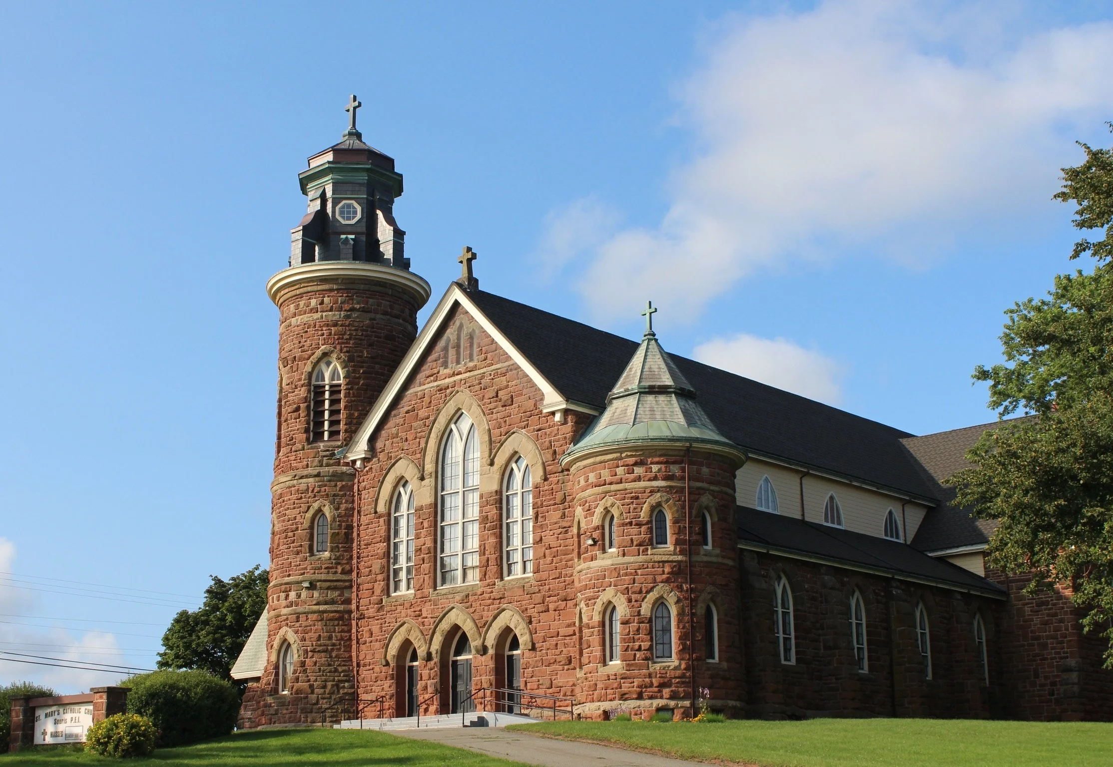 A historic church built with red stone, featuring tall arched windows, a prominent tower with a weather vane, and a cross at the top of the roof, set against a blue sky with clouds and surrounded by green grass and trees.