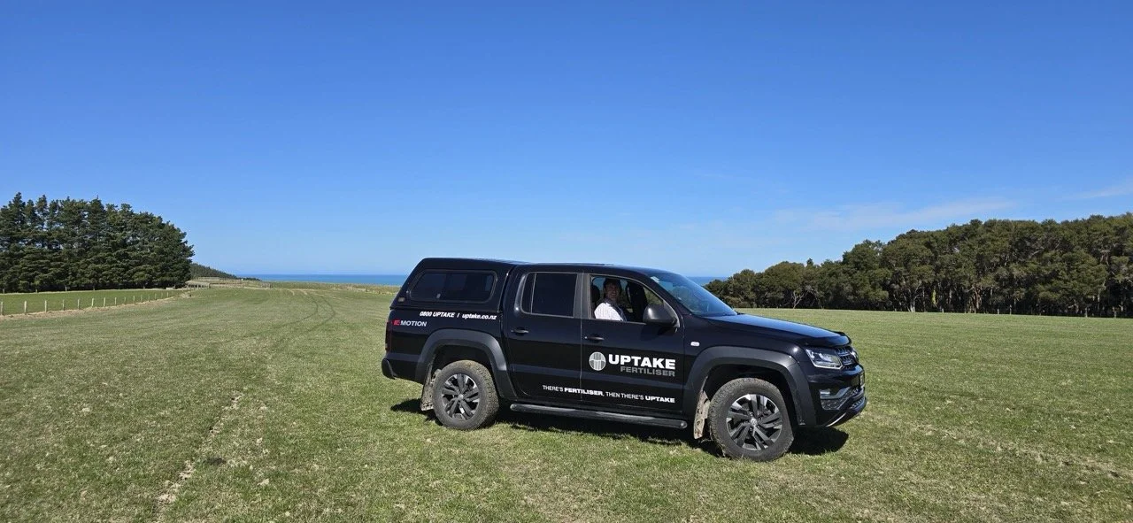 Black Uptake Fertilizer truck parked on a grassy field with a clear blue sky, trees in the background, and the ocean visible in the distance.
