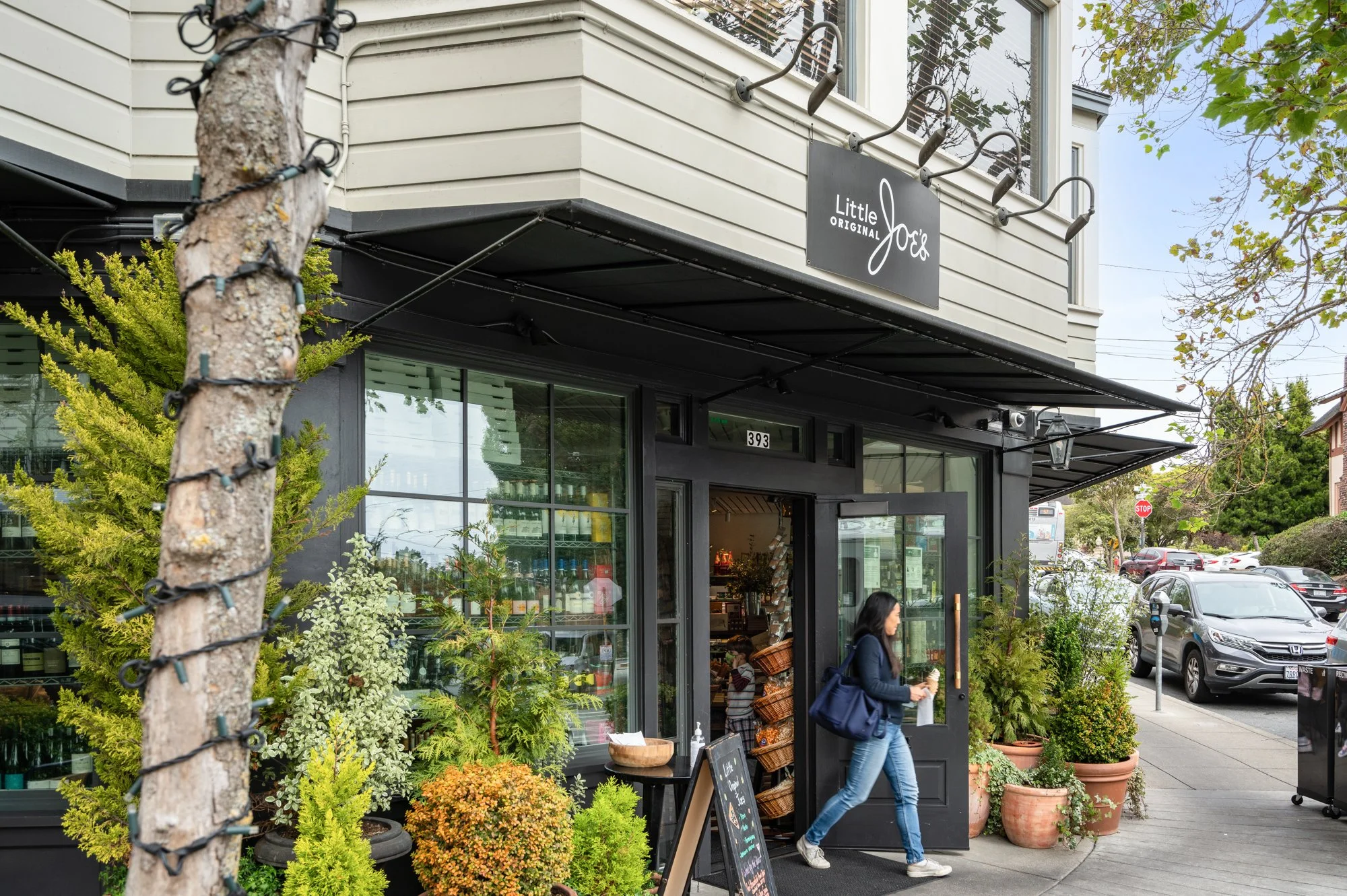 A woman with a dark jacket and jeans walks out of a storefront called Little Joe's, holding a drink and a bag, with potted plants outside, parked cars, and a street in the background.