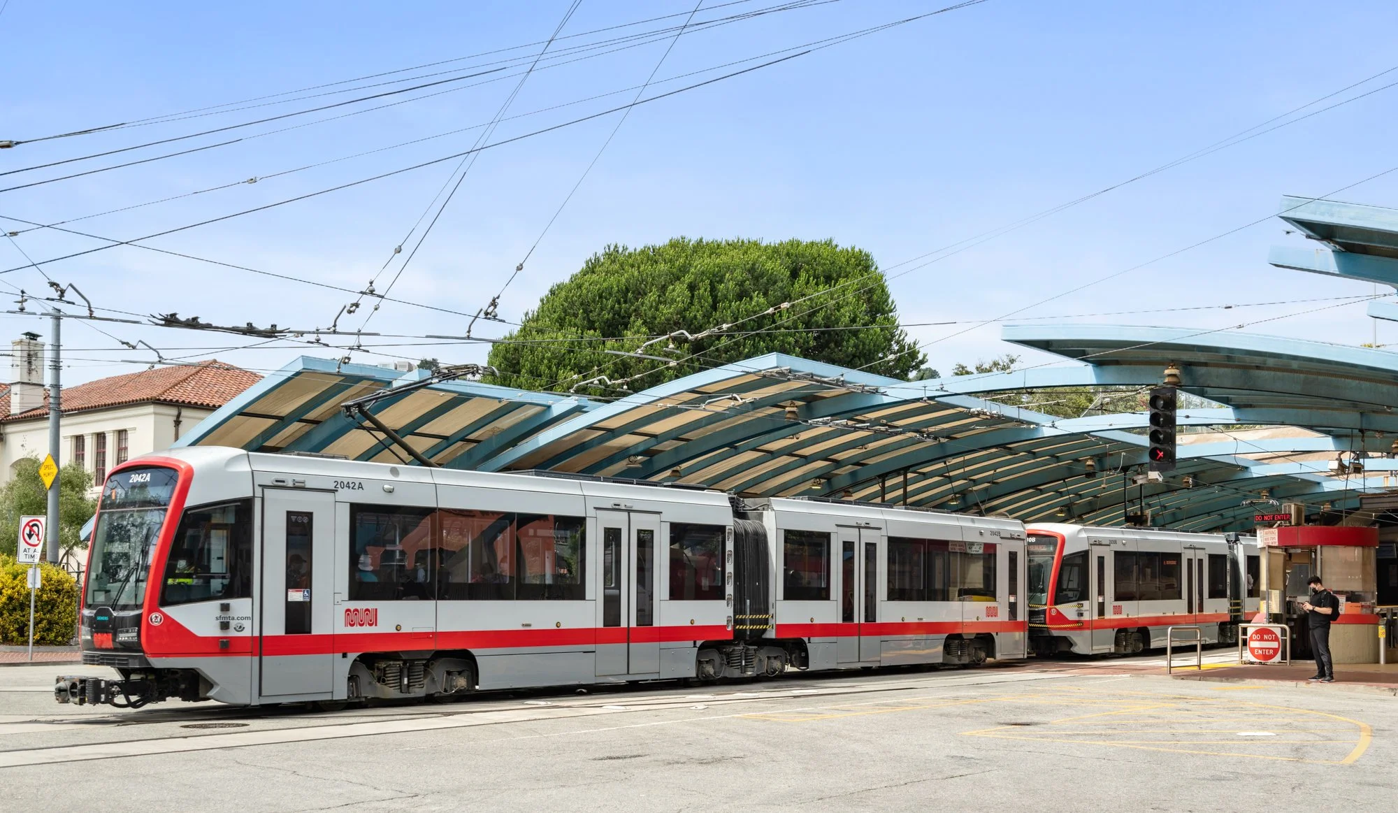 A modern light rail train at a station with a curved canopy, a person standing near the entry gate, and overhead power lines, in a sunny urban setting.