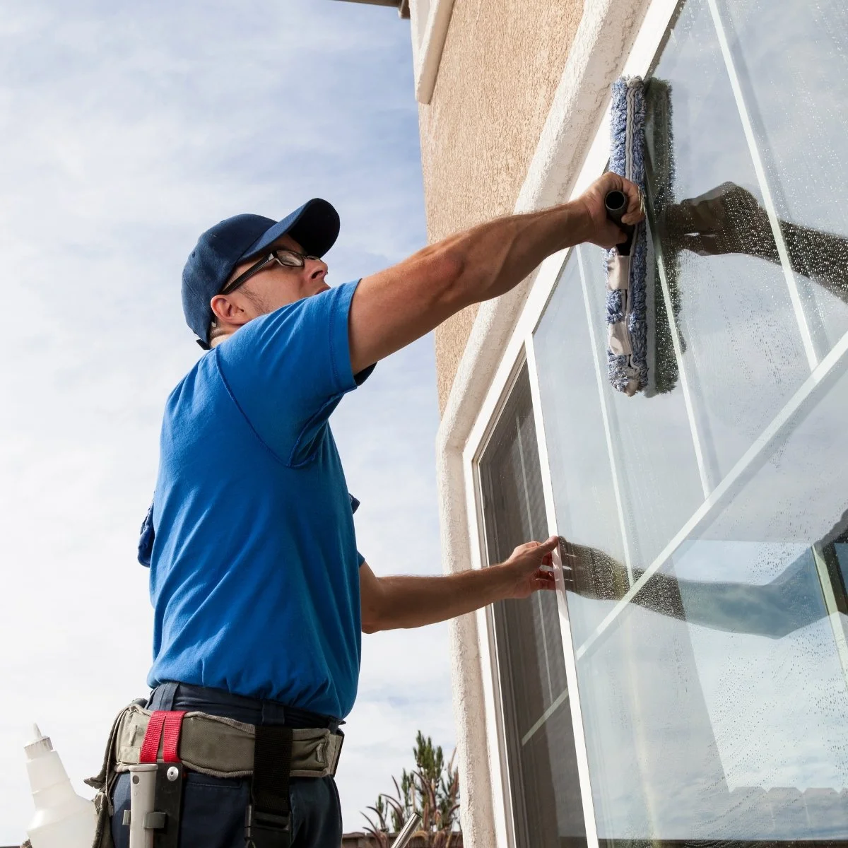A man cleaning a large glass window outside a house with a squeegee, wearing a blue shirt, glasses, and a baseball cap.