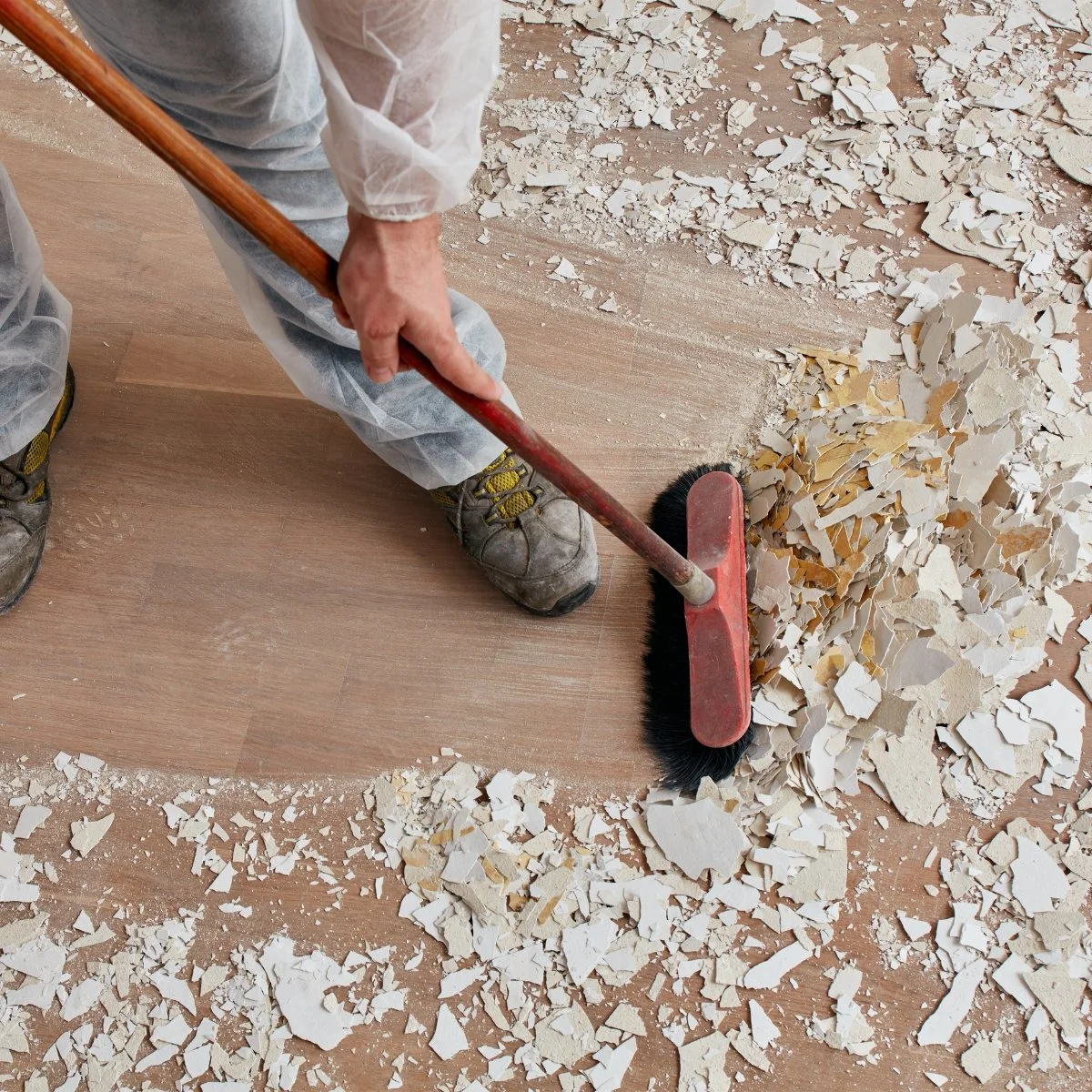 Person in work clothes using a dustpan and brush to sweep up torn pieces of drywall or plaster from a wooden floor.
