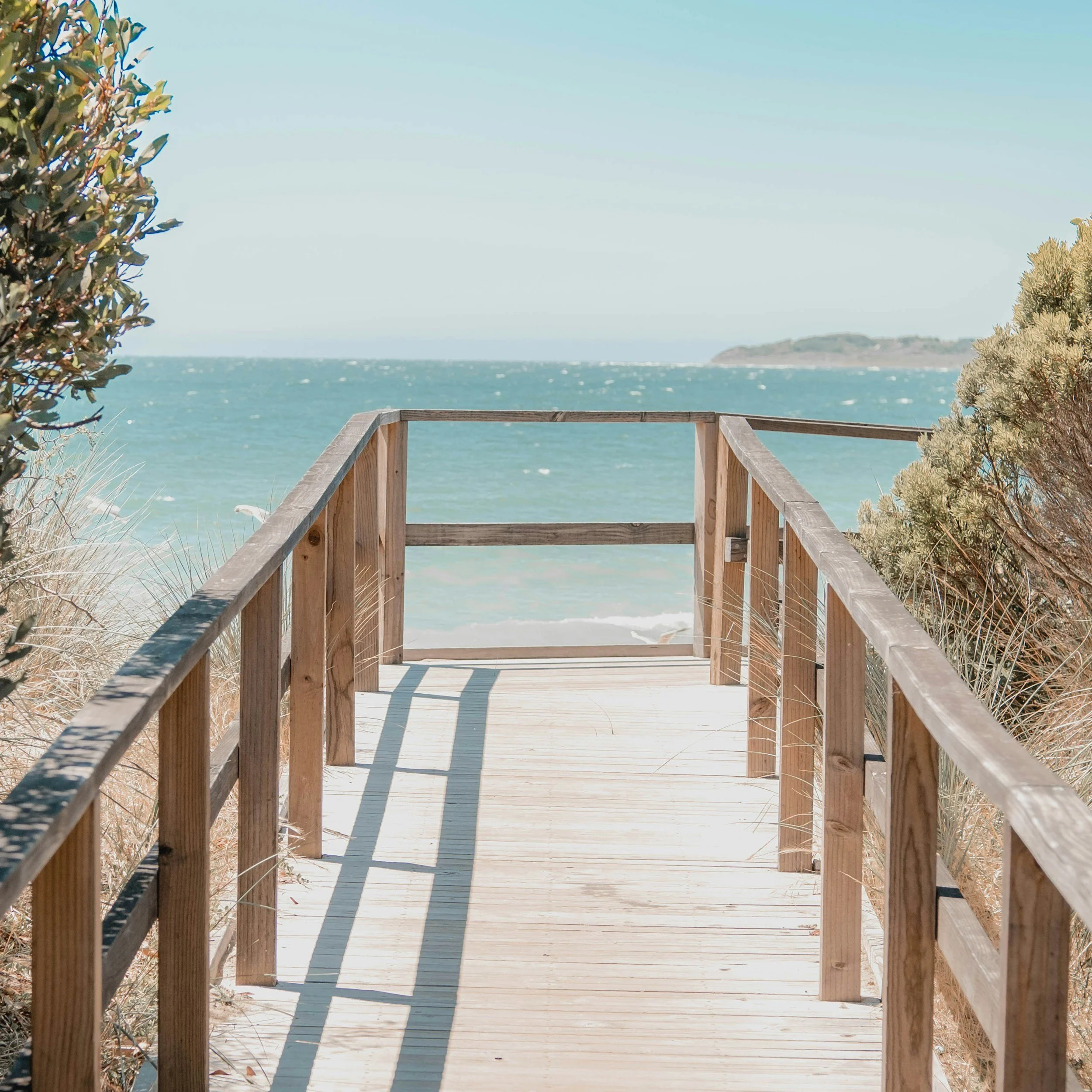 Wooden walkway leading to the beach, flanked by sand dunes and shrubbery, with the ocean and a distant land mass in the background.