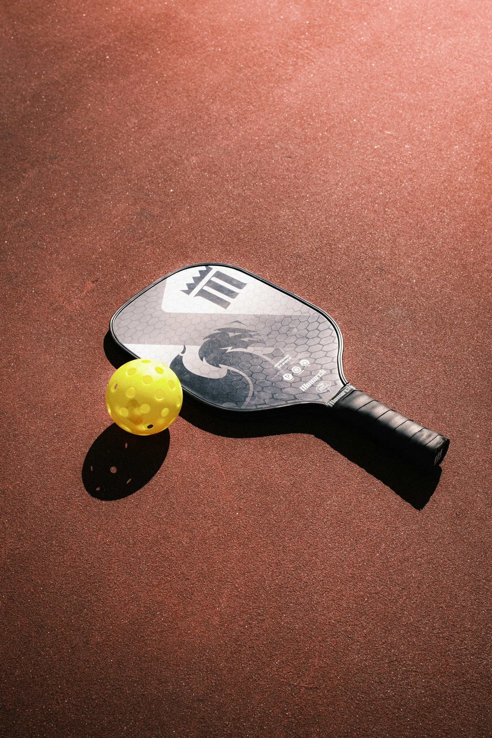 Padel racket with a dragon design and a yellow perforated tennis ball on a red outdoor court.