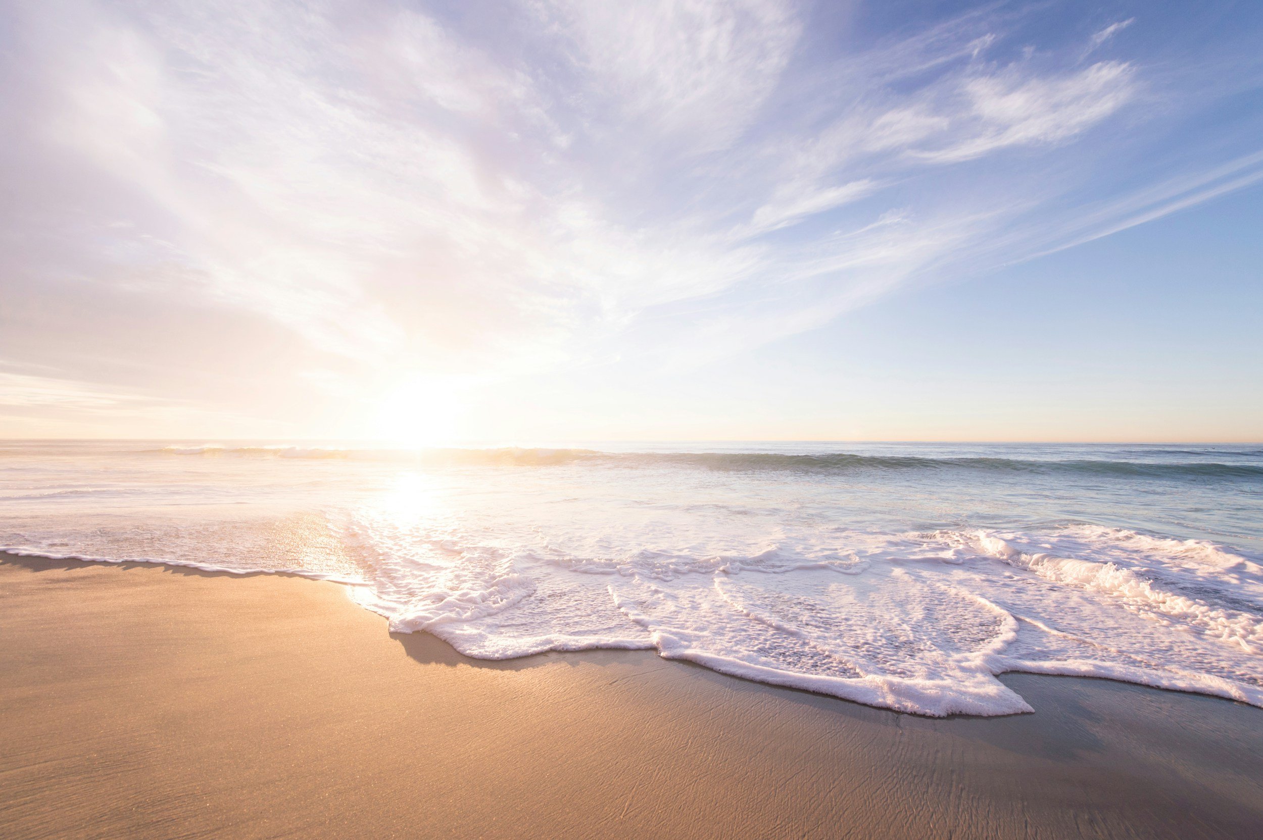 Sunrise over a calm ocean with gentle waves crashing onto a sandy beach, partly cloudy sky with pink and blue hues.