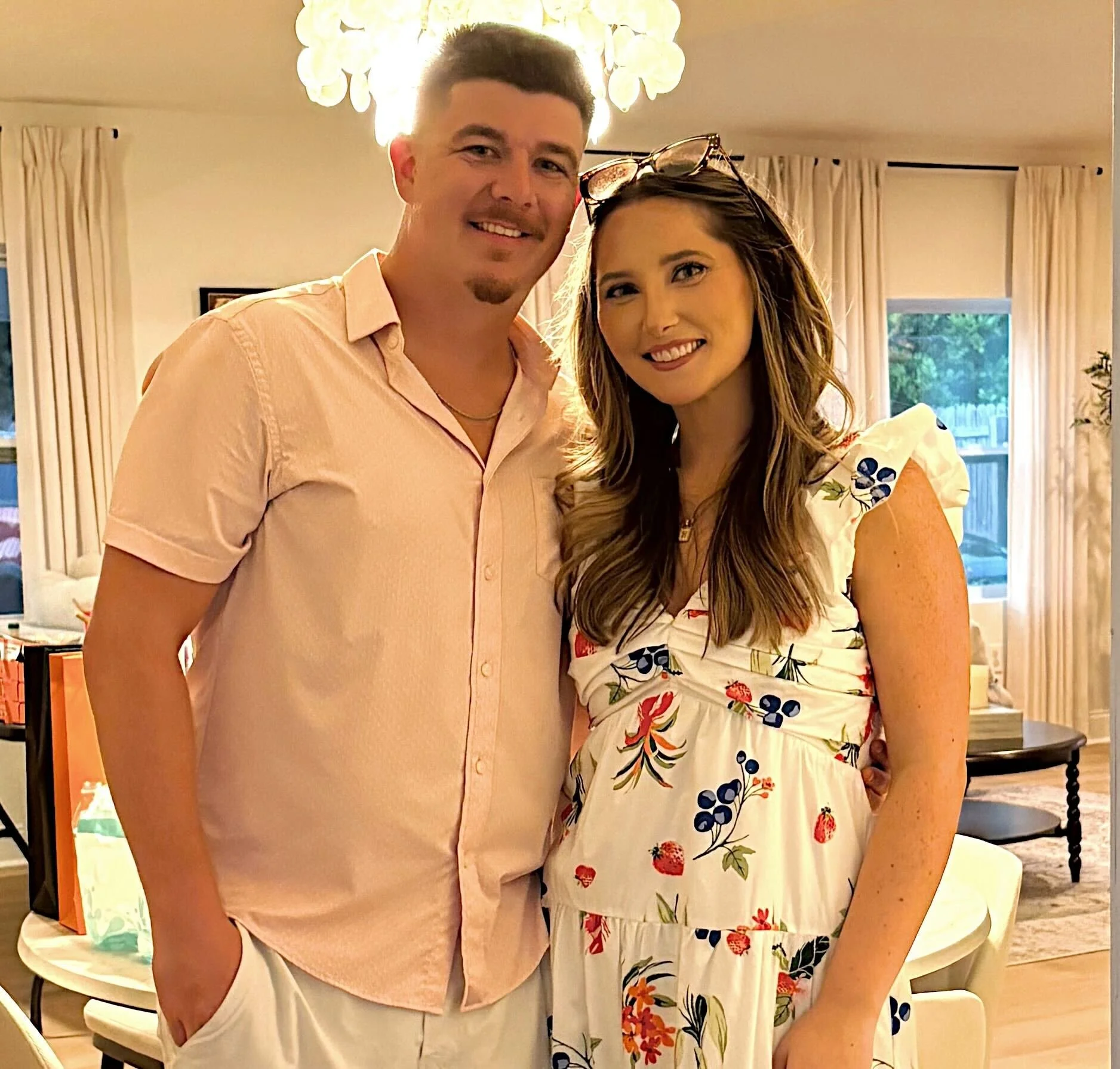 A man and woman standing close together indoors, smiling at the camera, with a chandelier overhead and a window with curtains behind them.