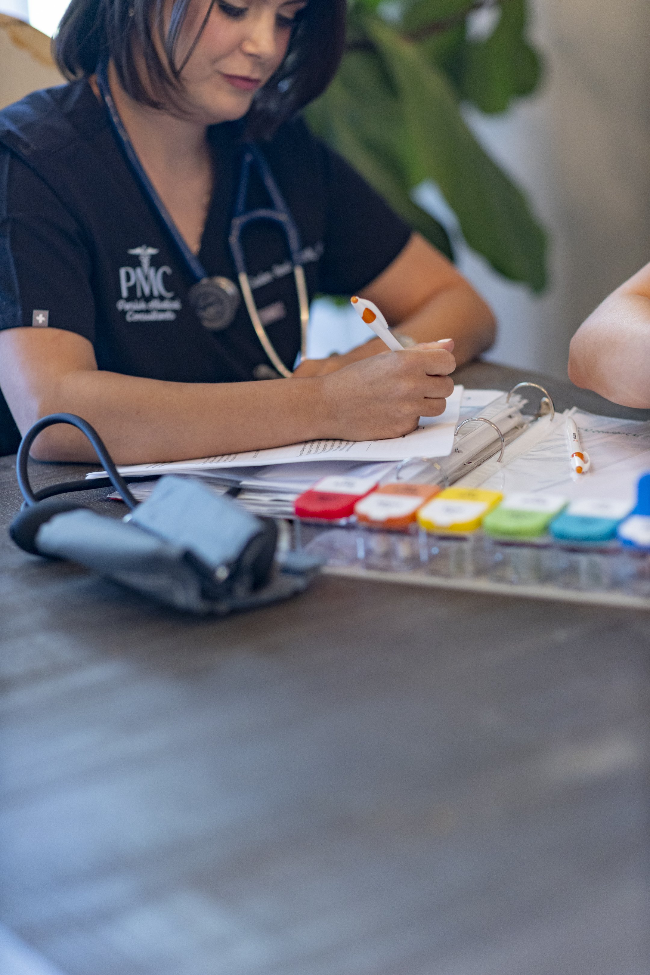 A healthcare professional, wearing a black polo with PNC logo and a stethoscope, is writing in a notebook on a table. Medical supplies, including a blood pressure cuff, blood test strips, and pens, are scattered on the table.