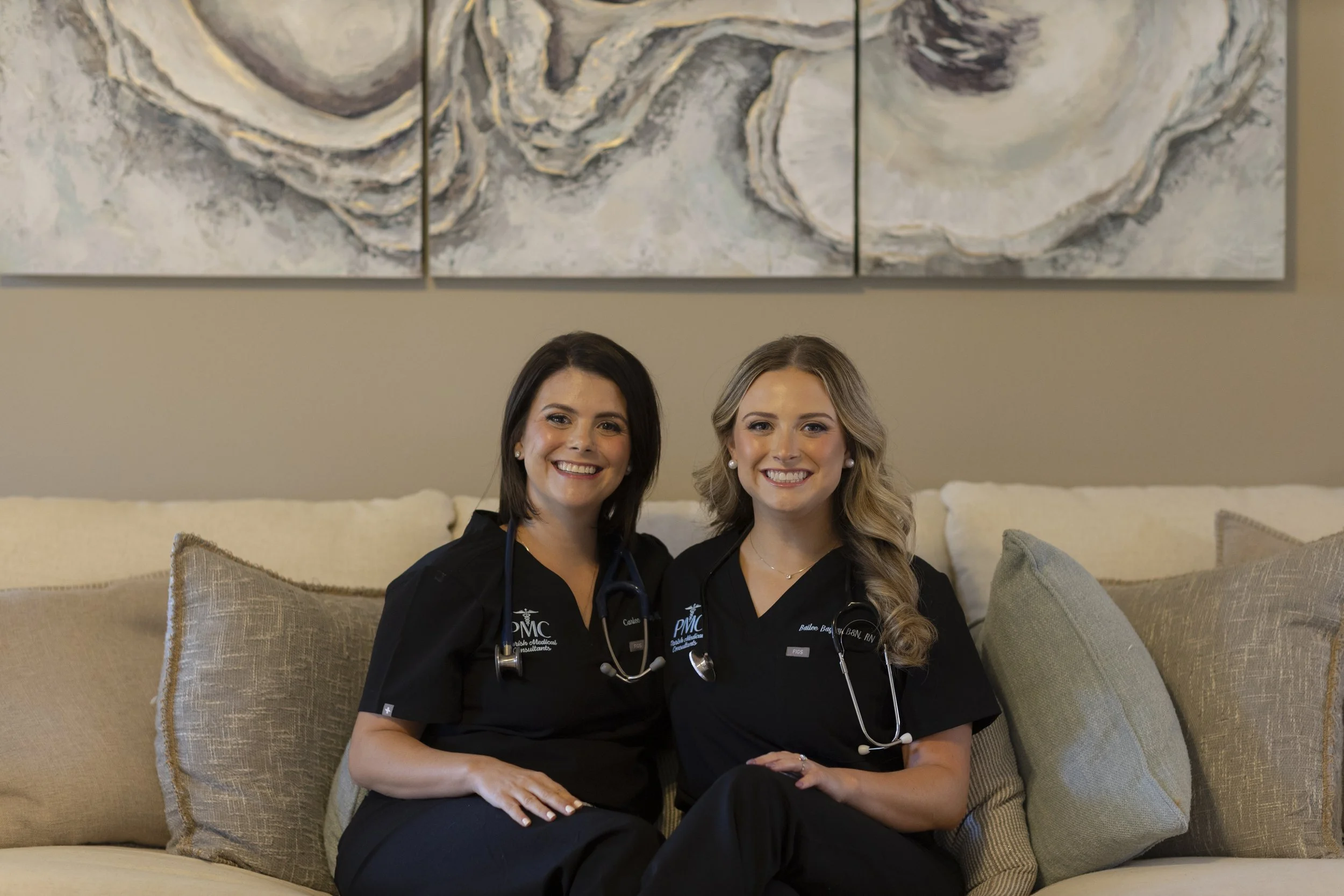 Two women smiling and sitting on a beige couch with pillows. Both are wearing black medical scrubs with stethoscopes around their necks, indicating they are healthcare professionals. Behind them is an abstract painting with light colors.