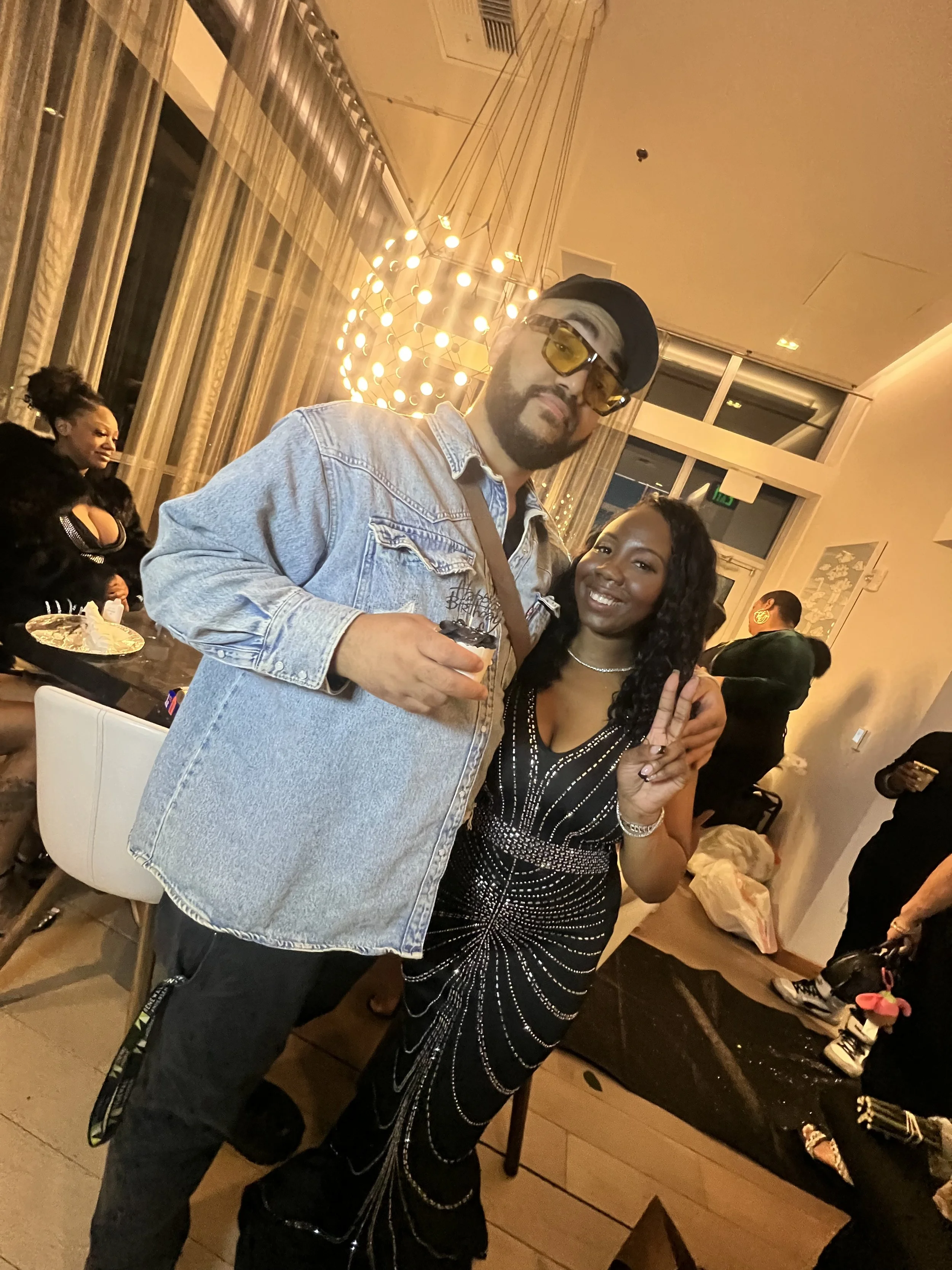 A man and a woman posing together at a party. The man is wearing glasses and a denim jacket, and the woman is smiling and making a peace sign. There are other people in the background and decorative lights from the ceiling.